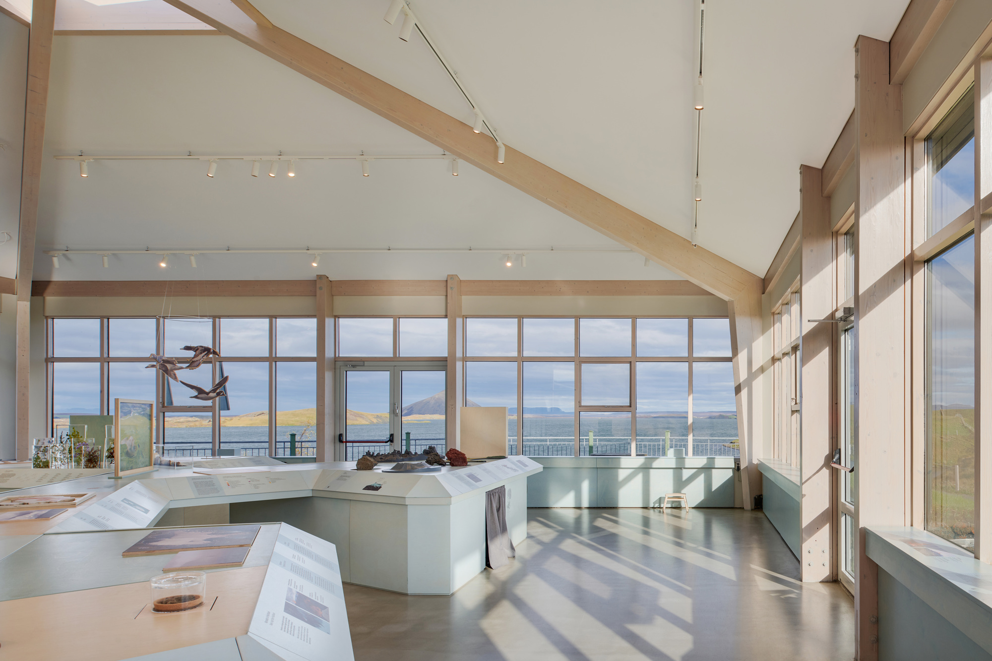 Interior of the exhibition room with timber roof beams, ribbon windows and angular display tables, lit by sun casting long window shadows