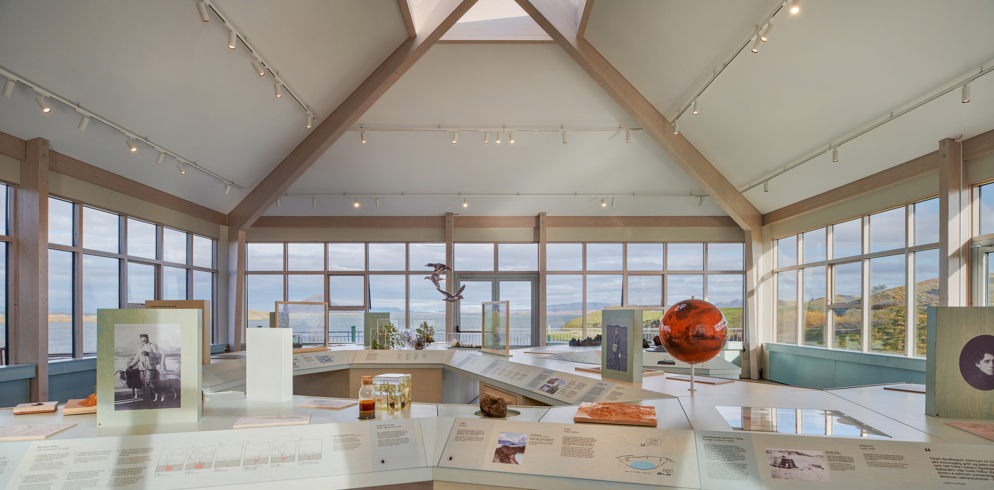 Exhibition hall with a timber-framed gabled ceiling, central skylight and display cases holding photographs, a red globe and a hanging bird model