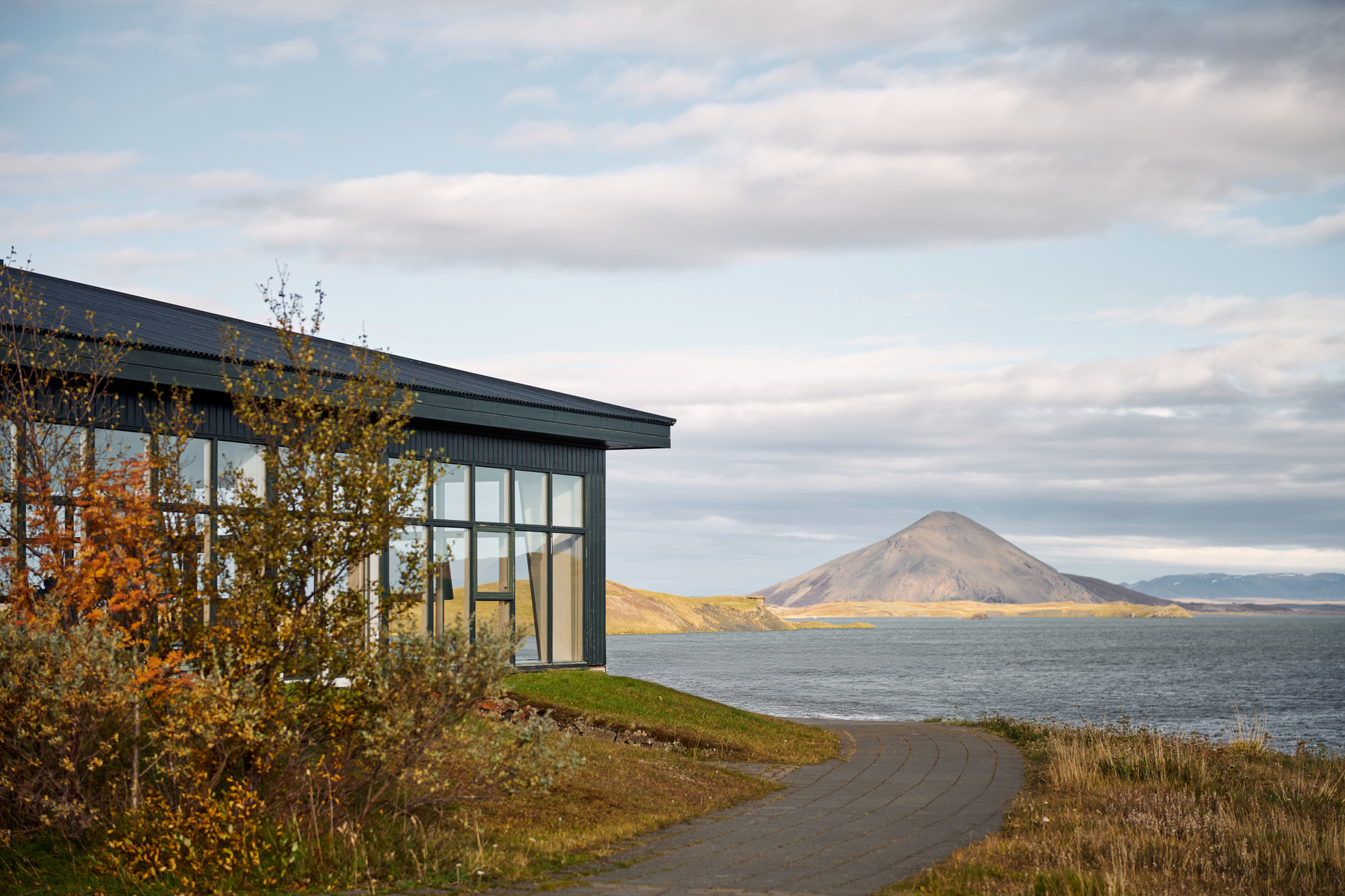 Dark timber-clad building with a corner glazing wall beside a curving paved path, facing a lake and conical peak in autumn