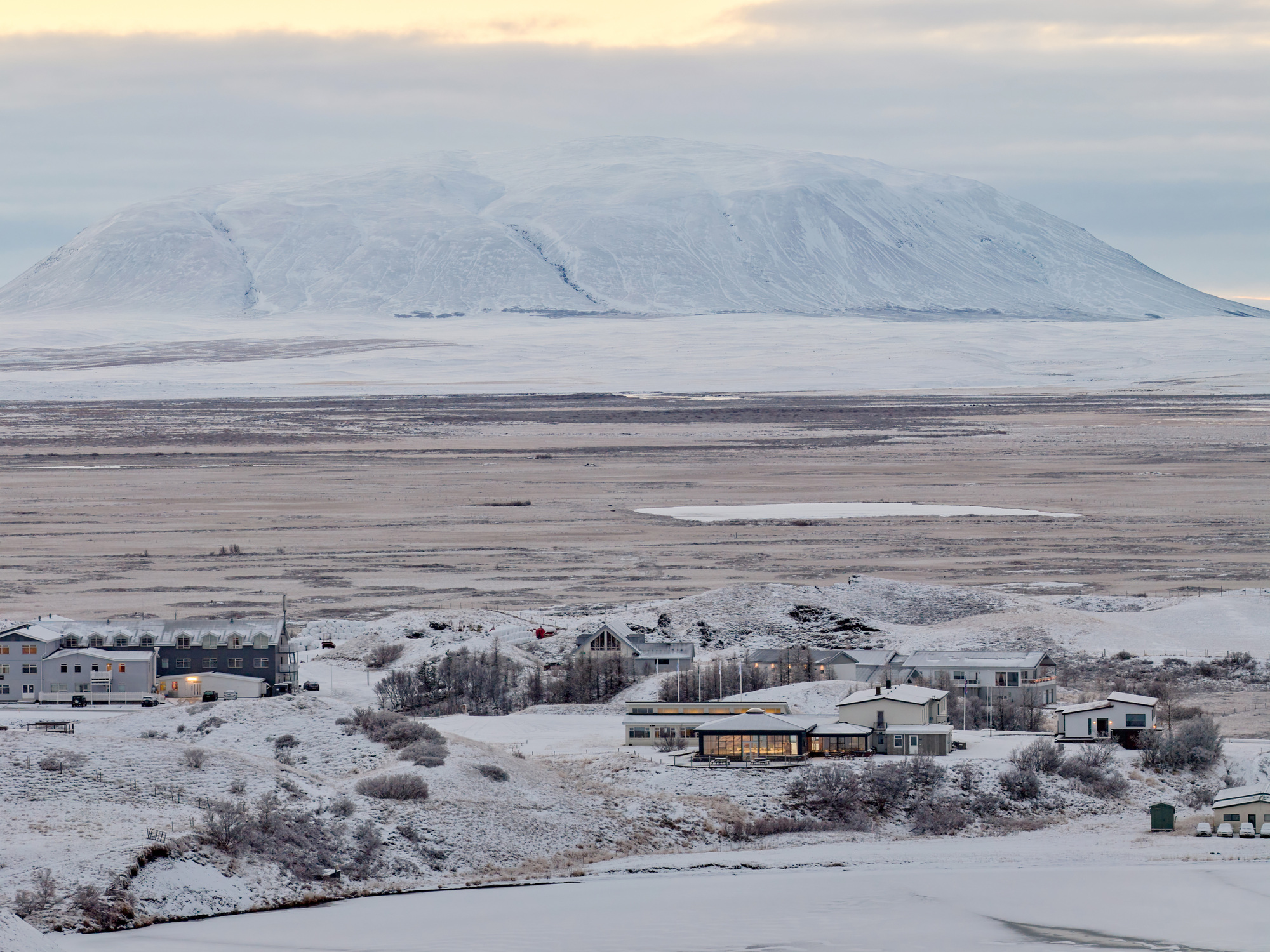 Distant view of a small cluster of buildings on snowy ground below a broad snow-covered mountain under a pale winter sky
