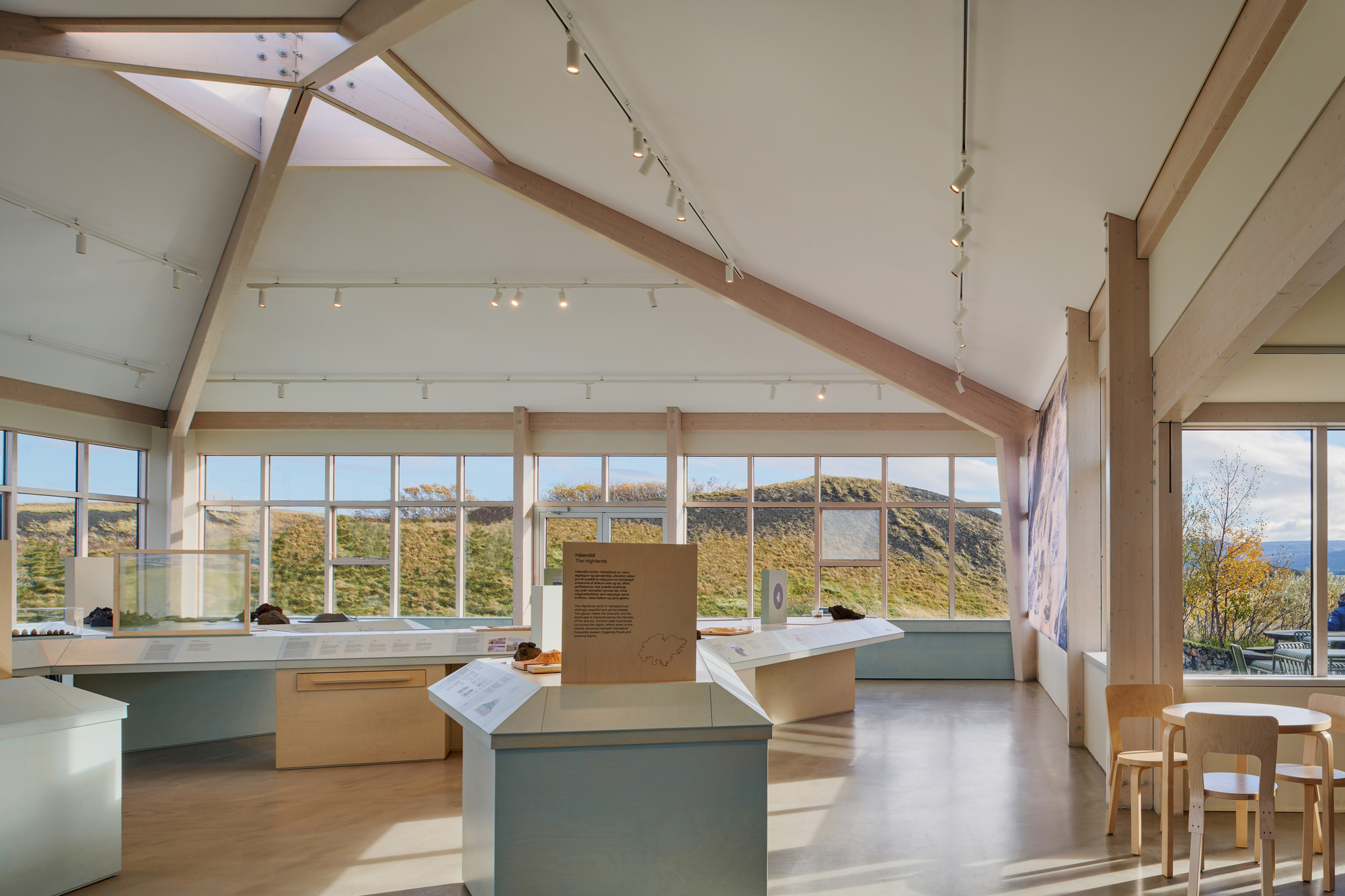 Exhibition room with pitched timber roof and glazed walls looking onto green hills, with small wooden chairs and a round table in autumn light