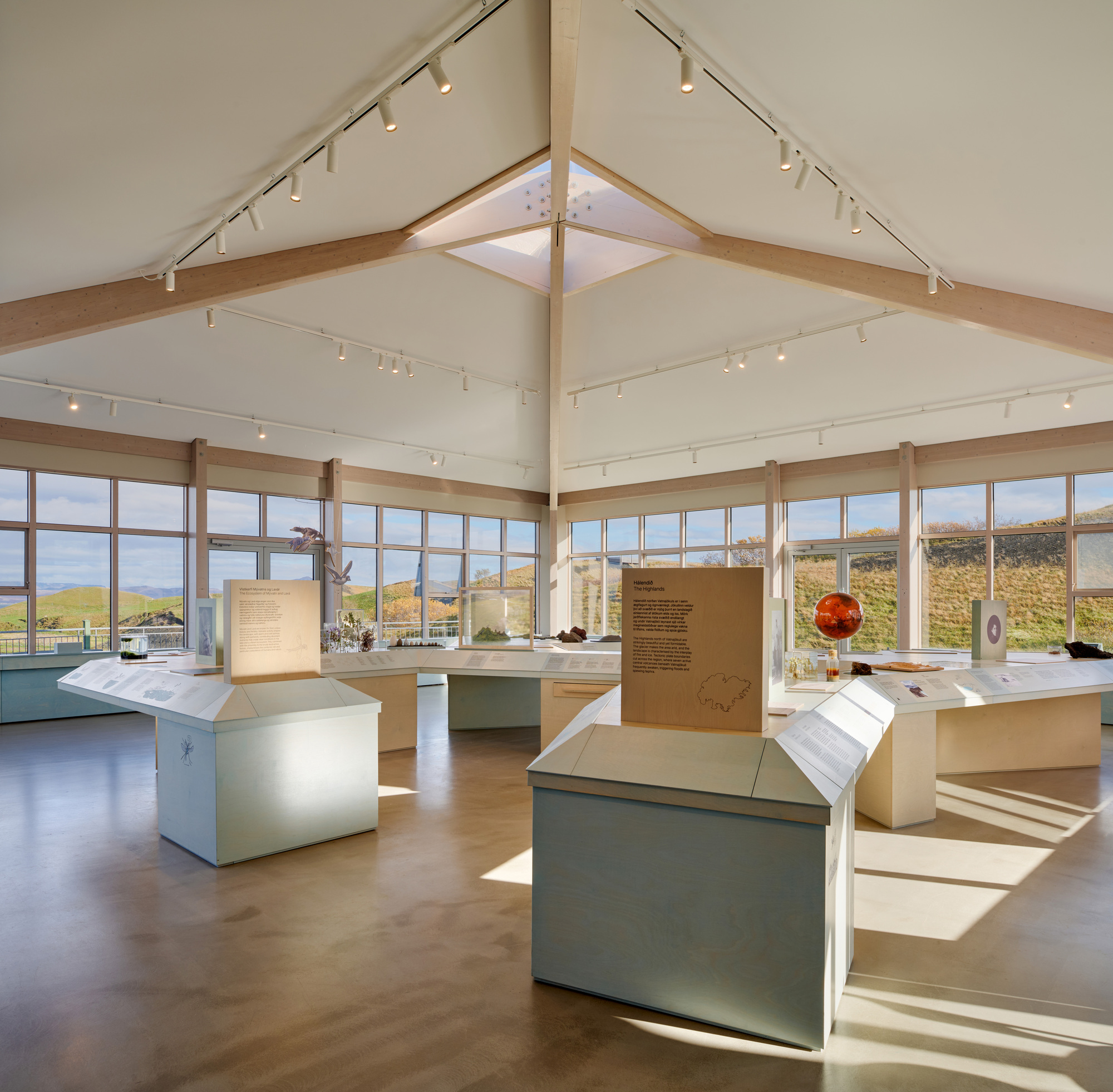 Display tables and a central roof skylight in the open exhibition room, with low sun casting bright patches across the floor