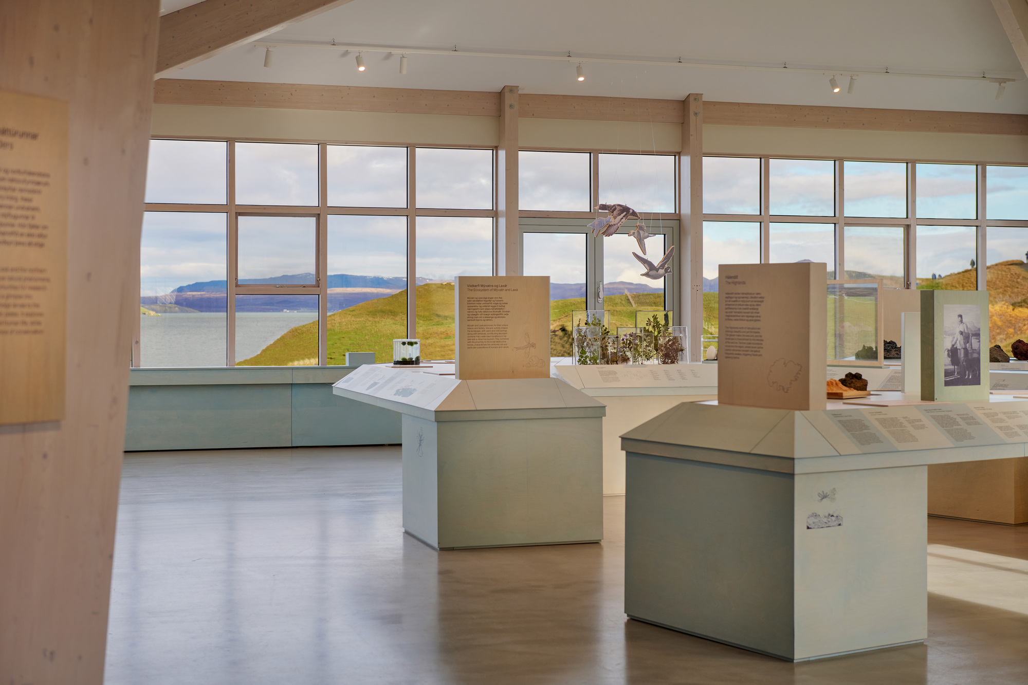Exhibition hall with pale green display plinths and a ribbon of windows framing a lake and grassy hills under cloud