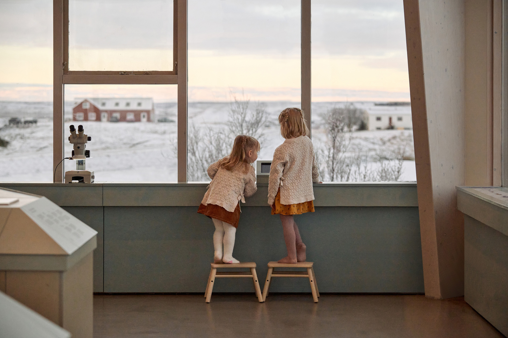 Two children on wooden step stools look through a tall window at a red farmhouse across snowy fields at dusk