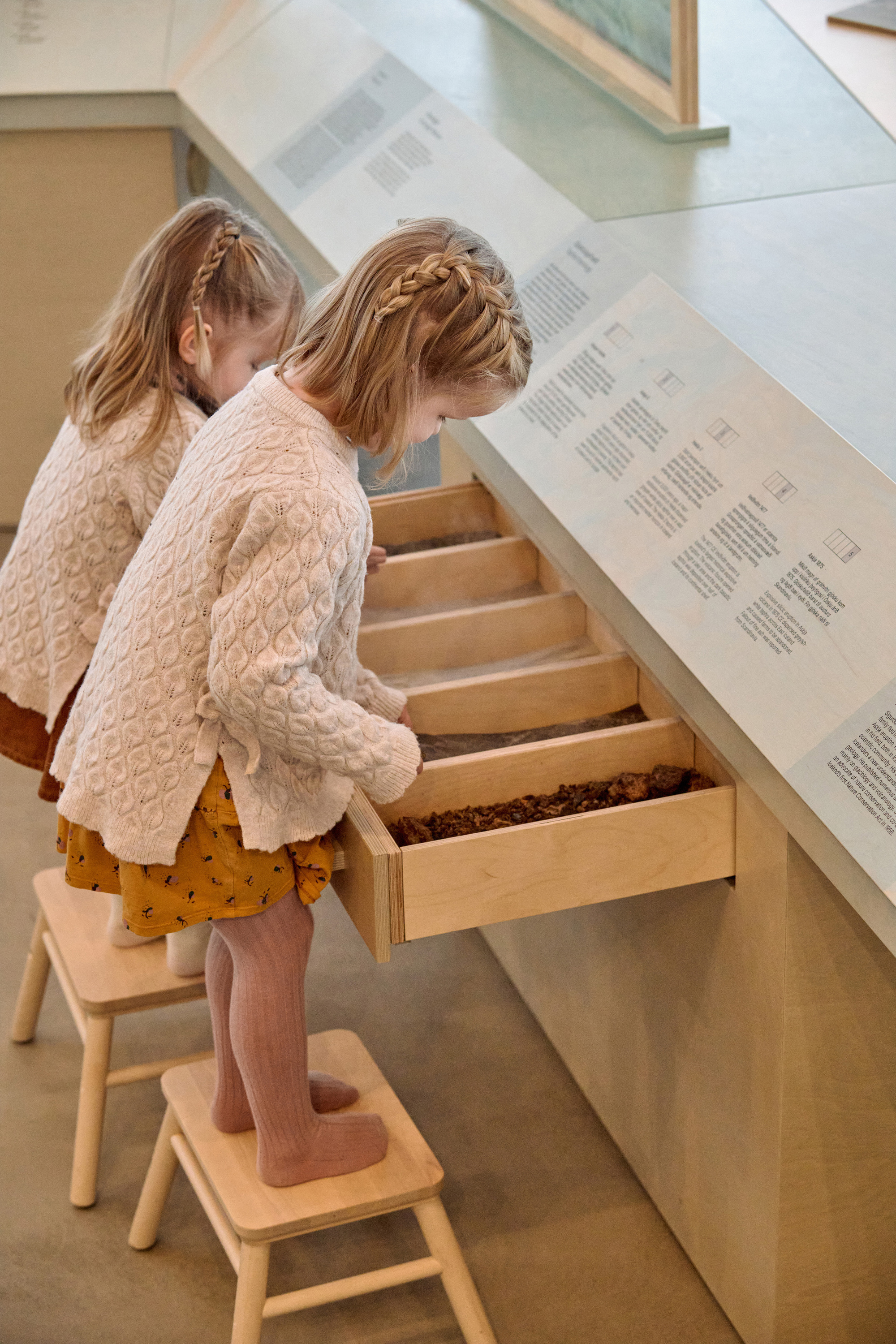 Child in a knit cardigan pulls open a wooden drawer of soil samples beneath a sloped display top with printed labels