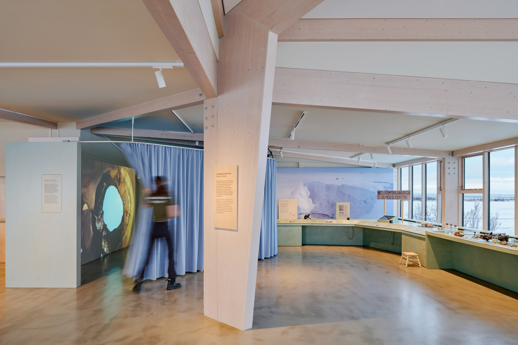 Gallery interior with a tilted timber column, blue curtain and a blurred visitor walking past a projected image
