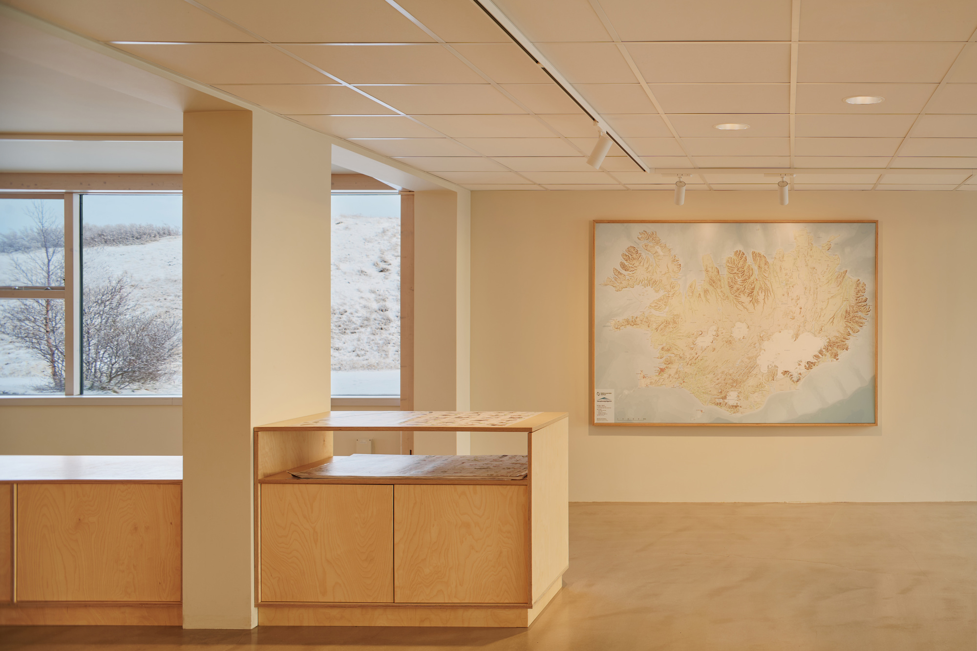 Framed relief map of an island on a cream wall beside a birch plywood cabinet and a snowy window view