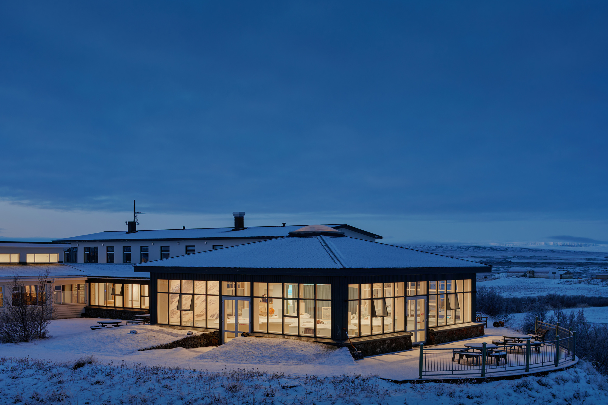 Glazed pavilion with a hipped roof glowing at dusk, a low wing behind it and snow across the surrounding ground