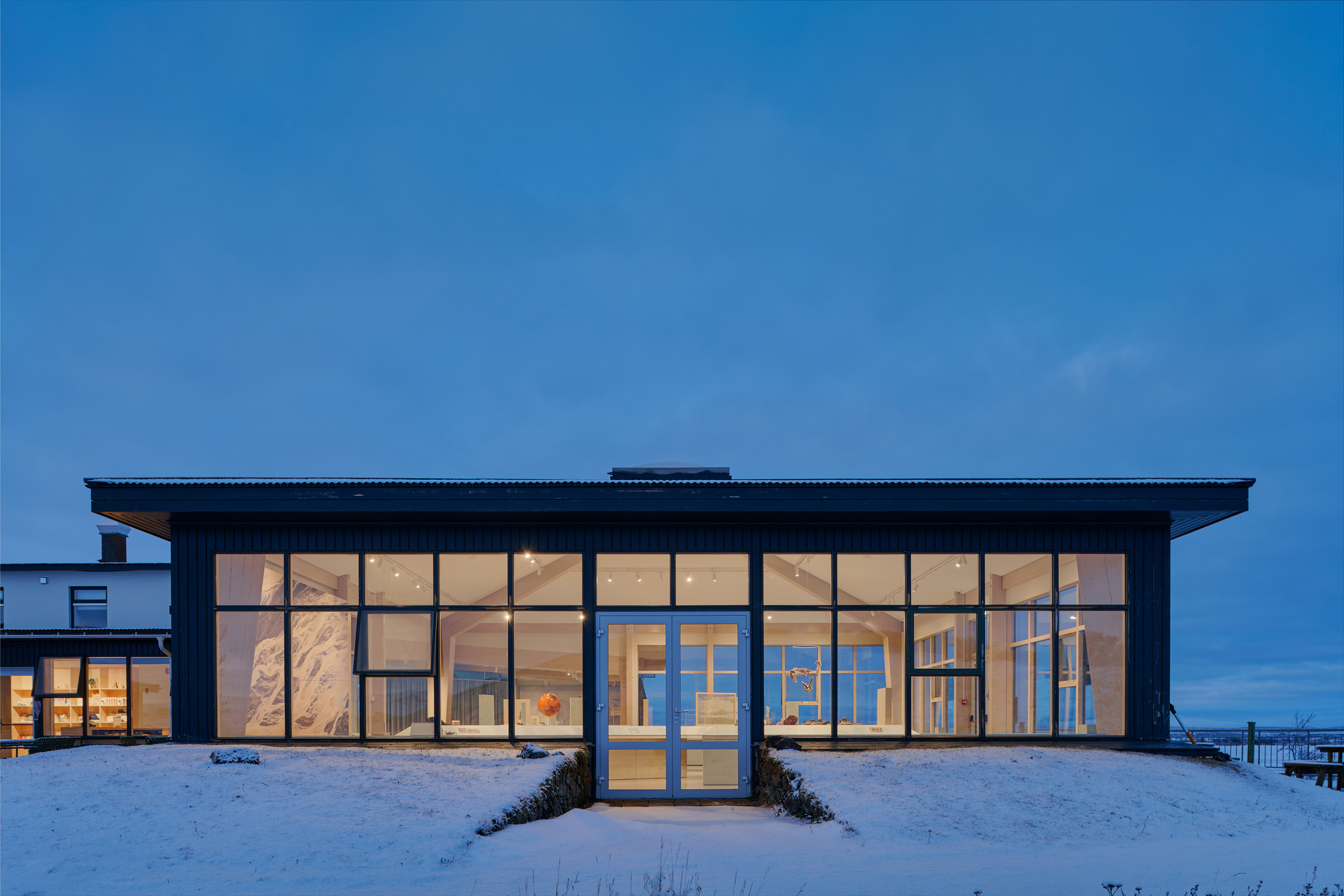 Front elevation of the dark timber-clad pavilion with full-height glazing and a central double door, lit warmly under a blue twilight sky