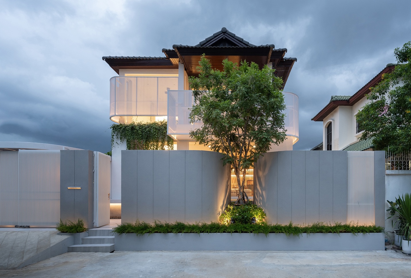 Street facade with curved grey panel wall, a single tree and translucent balcony screens glowing under a stormy dusk sky