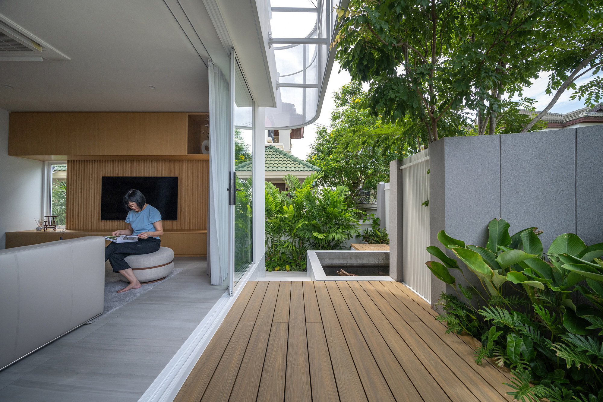 Timber deck beside an open sliding glass wall, with a woman reading in the living room and a small pond outside