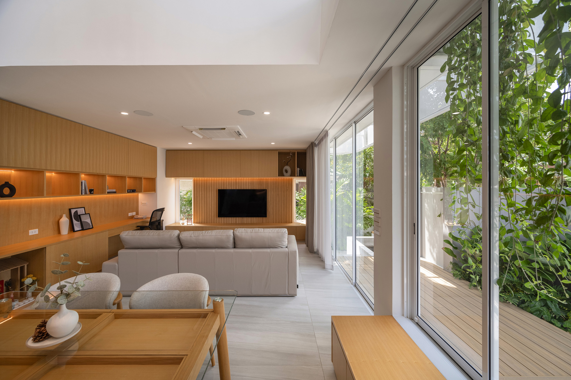 Living area with oak joinery, grey sofa and floor-to-ceiling glass opening onto a deck and climbing vines in daylight
