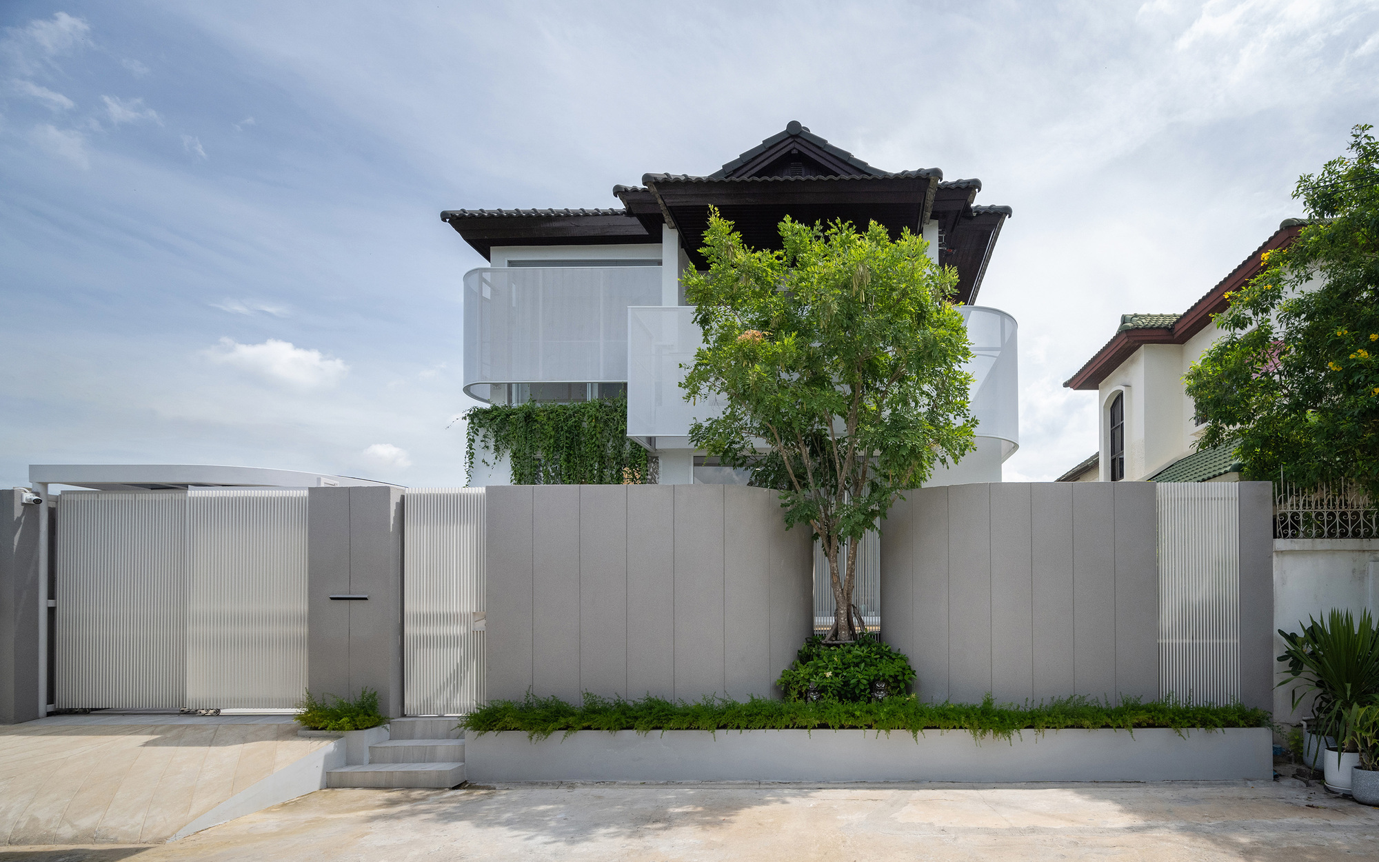 Front elevation with fluted gate panels, a planted bed and a small tree beneath curved balconies on a partly cloudy day