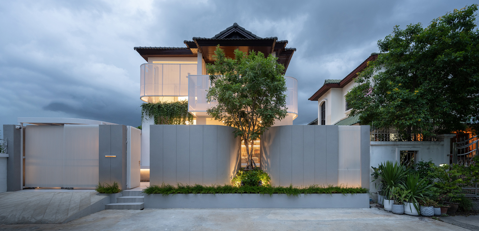 Street facade with gray panel walls, a central tree and translucent curved balconies under a heavy dusk sky