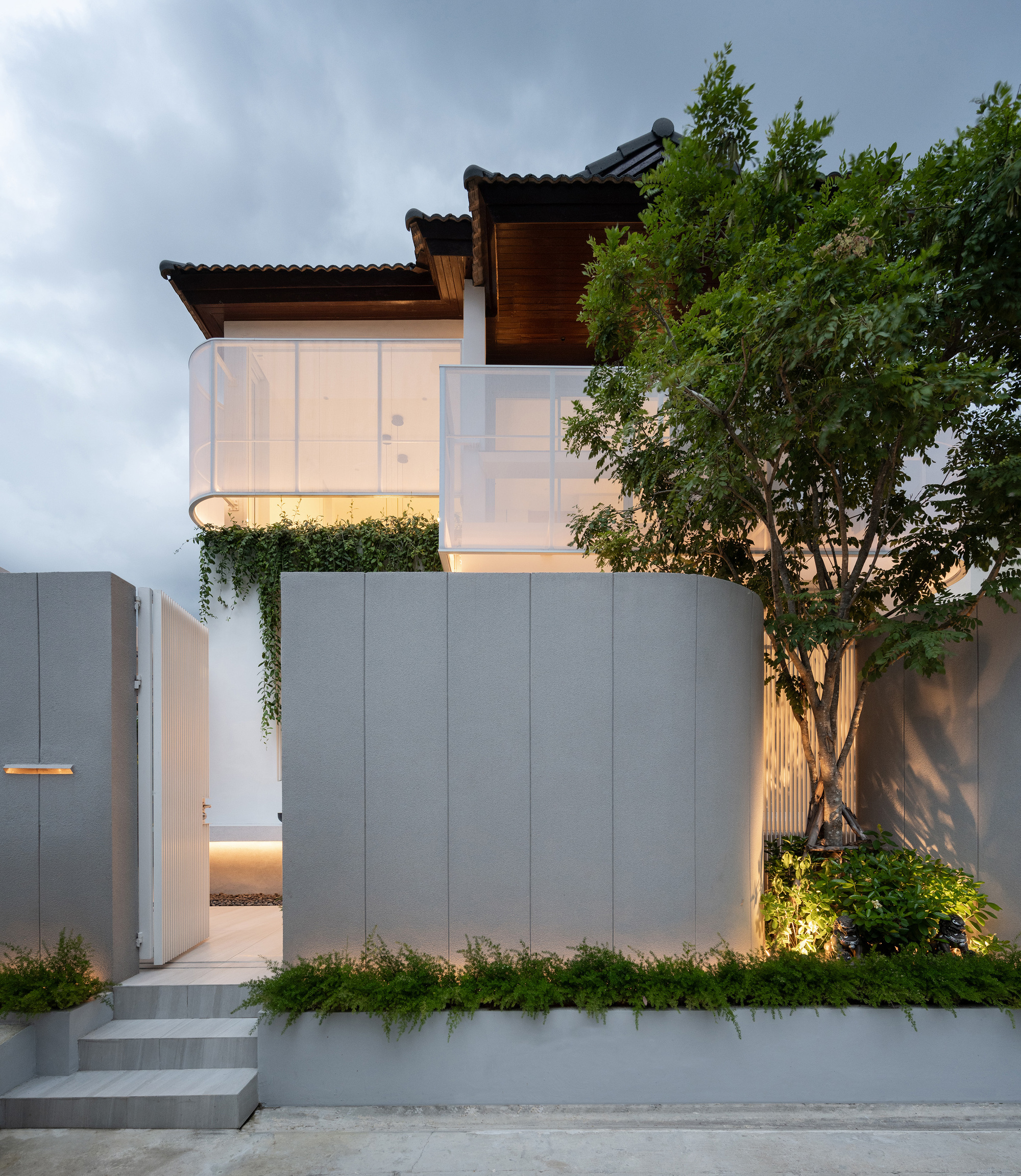 Curved gray panel wall and open gate below white balcony screens, with a leafy tree lit at dusk