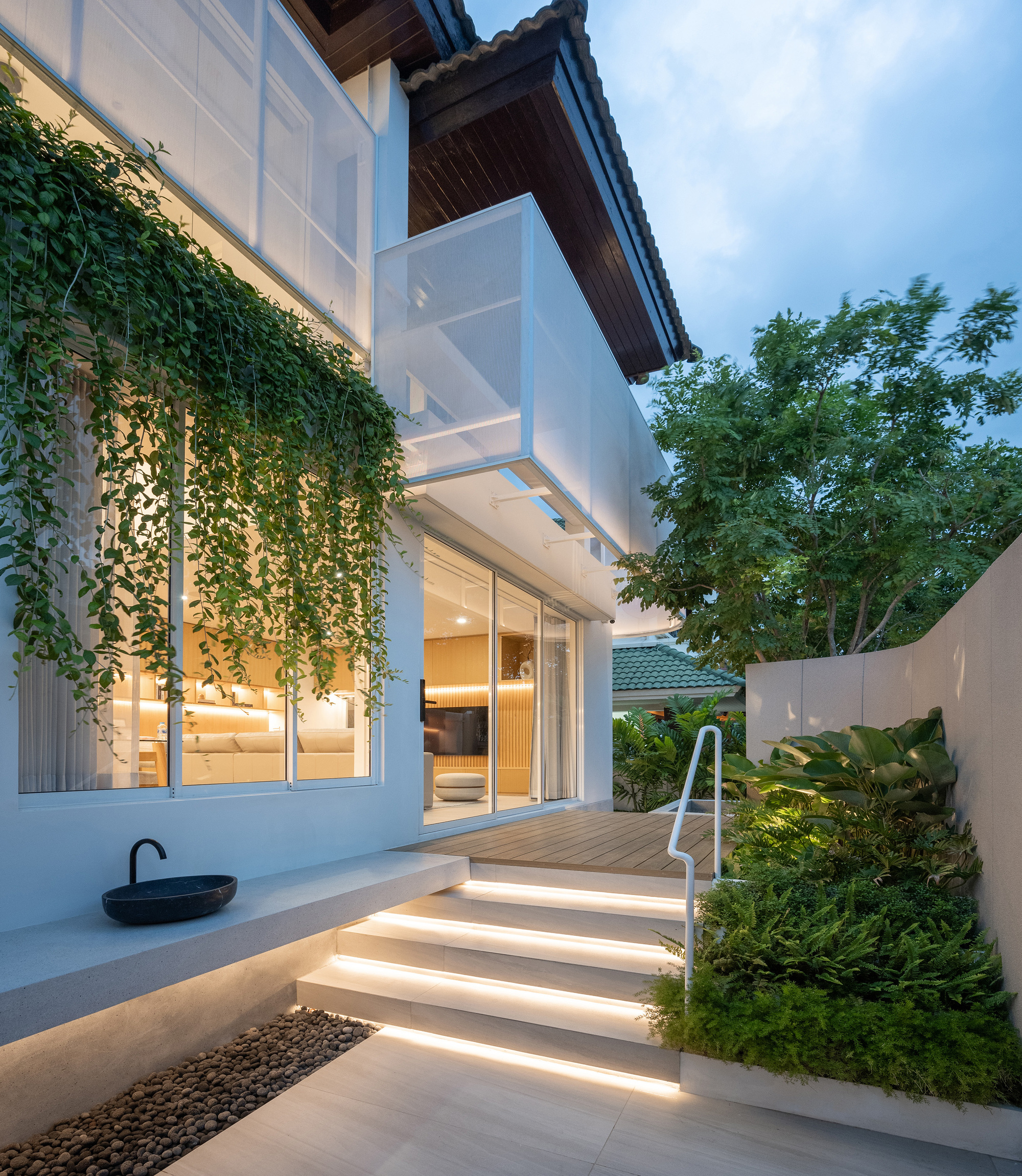 Side entrance with lit stone steps, a black basin and hanging vines beside a glazed living room at dusk