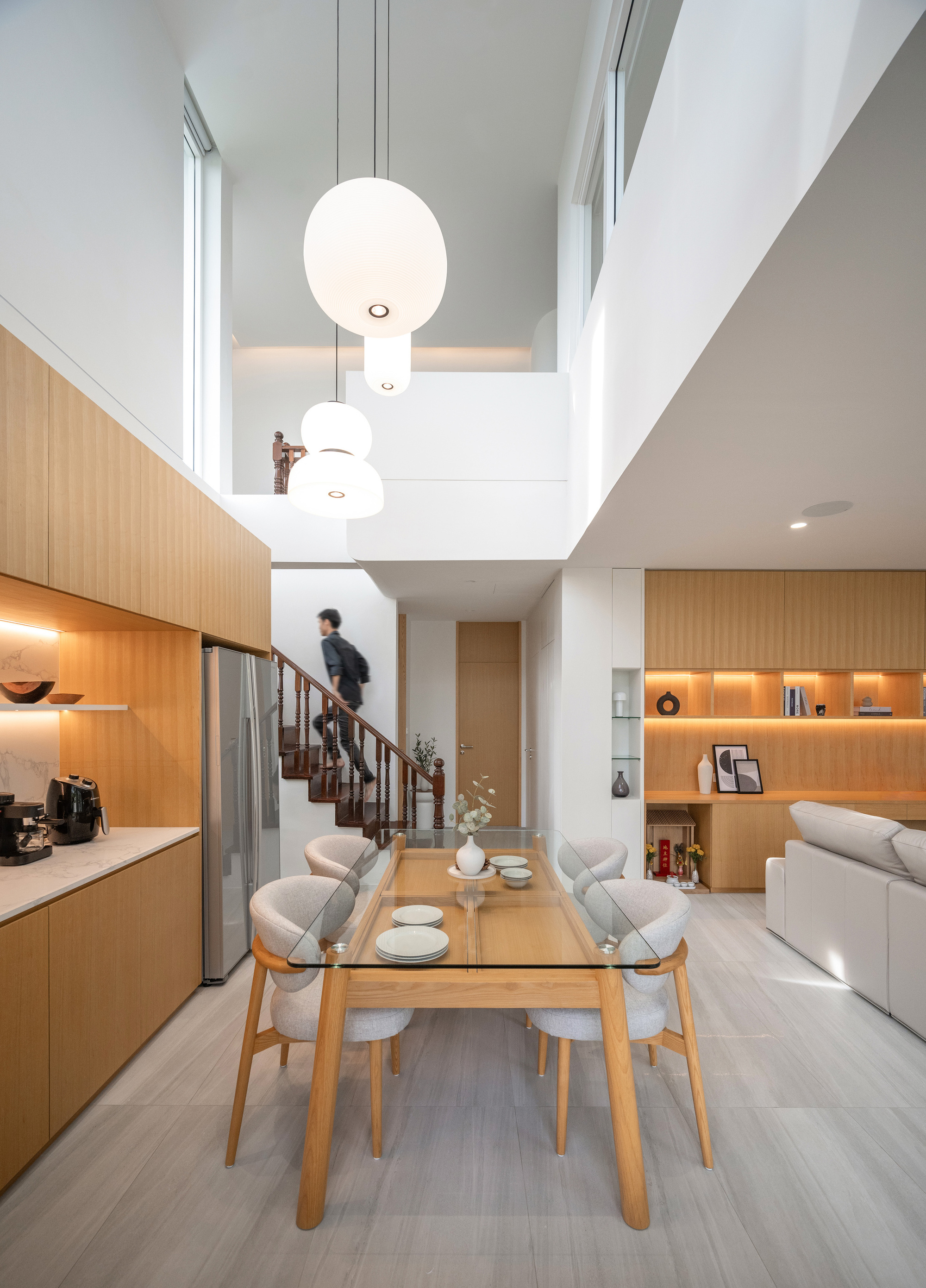 Double-height dining area with glass table, globe pendants and a person climbing the timber staircase behind