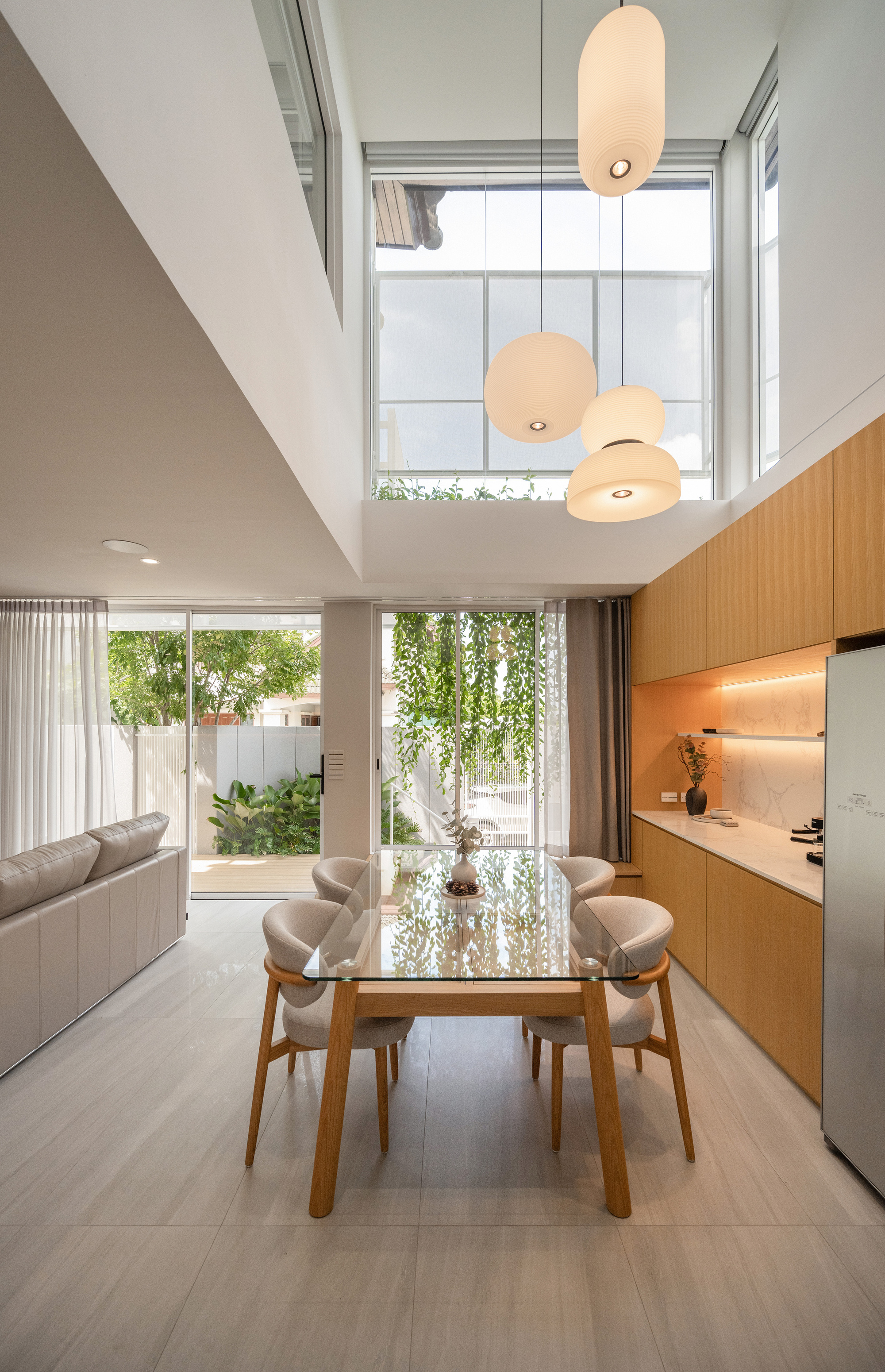 Dining table and pendant lamps in a double-height room, with vines and a garden visible through sliding glass doors