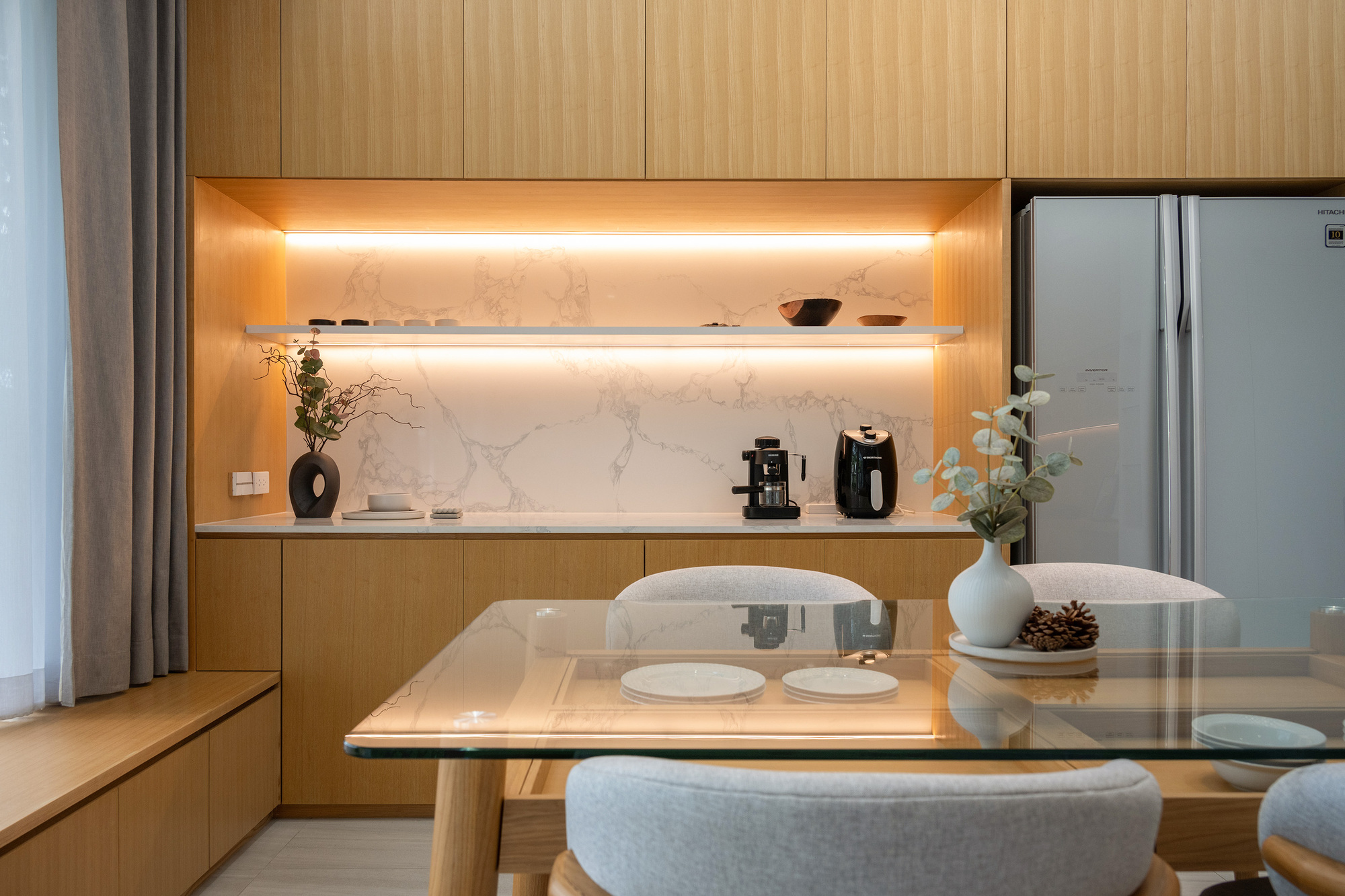 Kitchen niche with marble backsplash, lit floating shelf and oak cabinetry behind the glass dining table in soft daylight