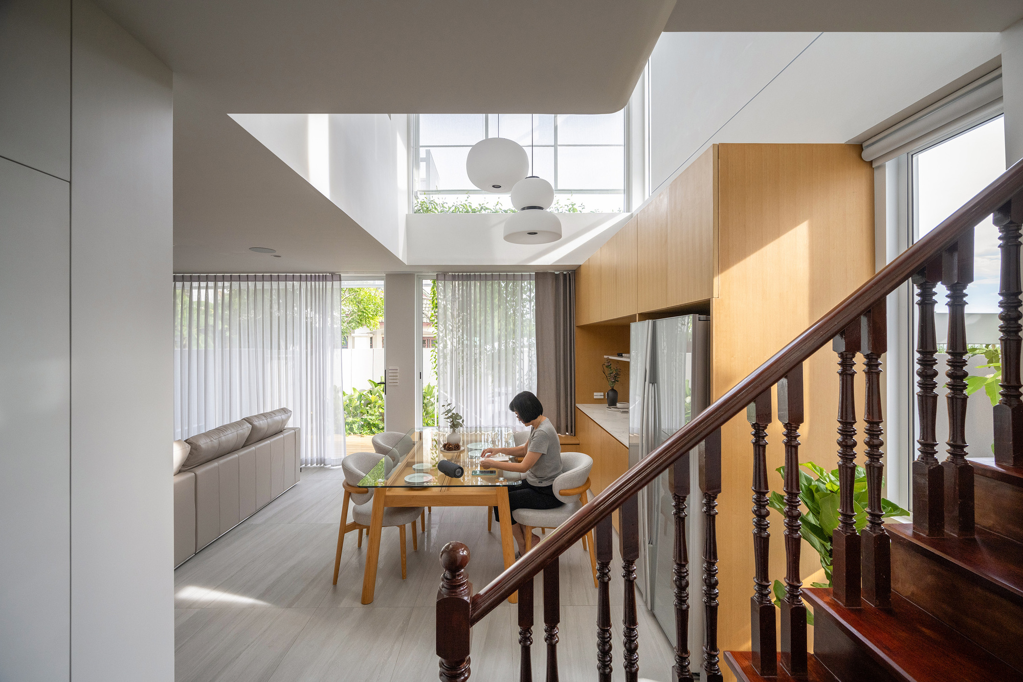 Double-height dining area with a turned timber stair balustrade in the foreground and a woman working at a wooden table in daylight