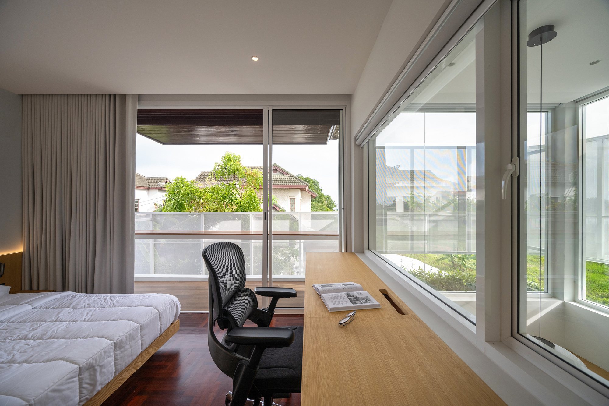 Bedroom corner with a built-in oak desk, mesh task chair and sliding glass door opening onto a balcony with trees beyond