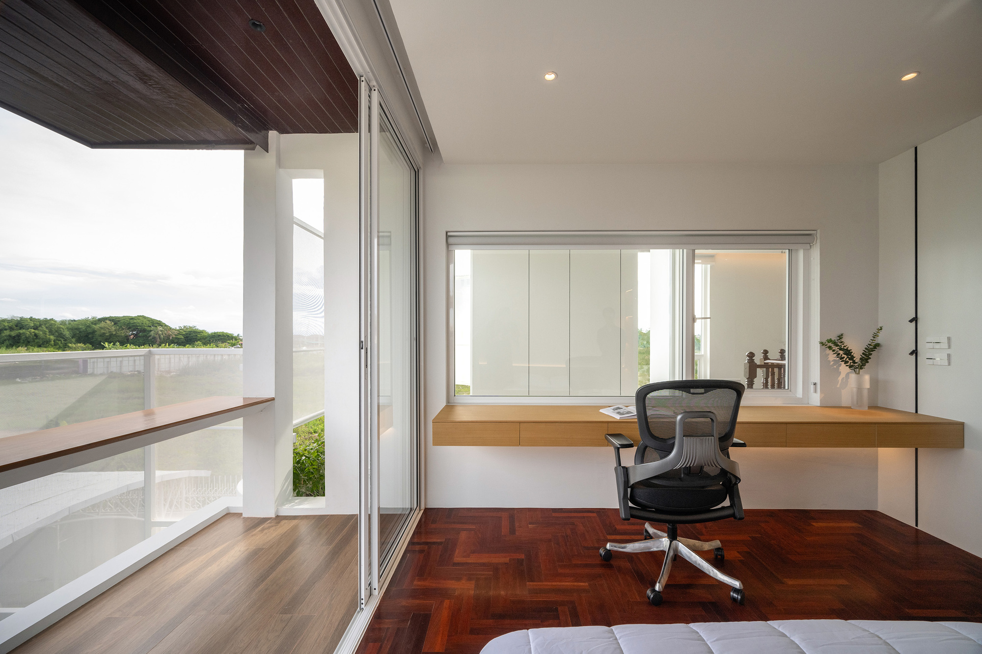 Floating oak desk under an interior window, with a sliding glass door to a balcony ledge above green fields under overcast sky
