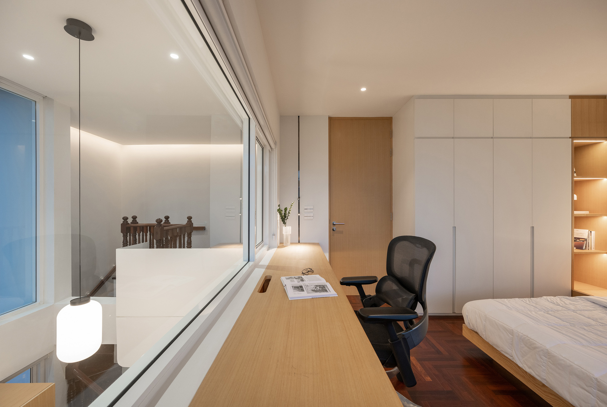 Timber desk running along an interior glass wall beside a bed and white wardrobes, lit by warm dusk light