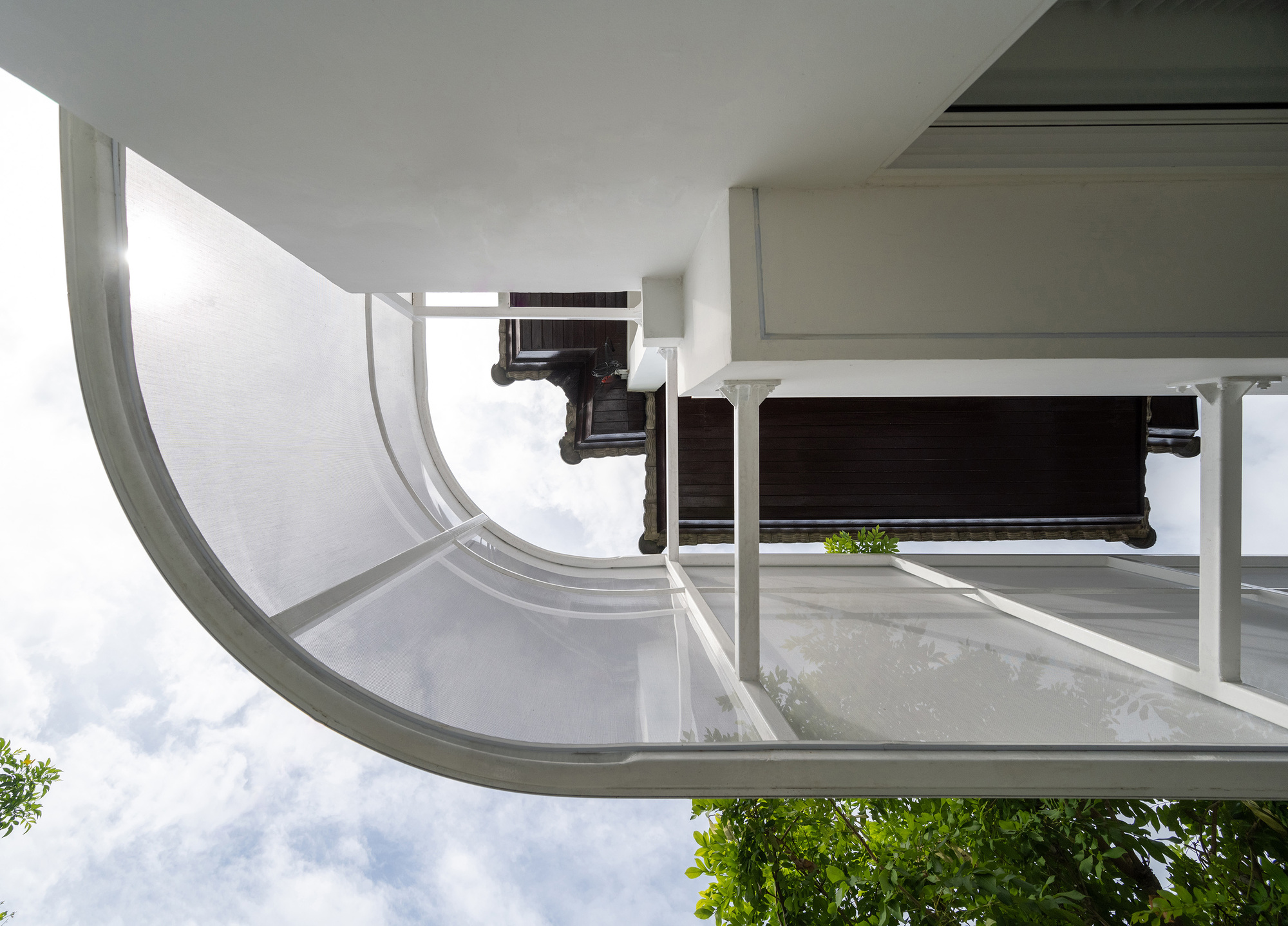 Upward view of a curved white mesh balcony edge against a cloudy sky with leafy branches below