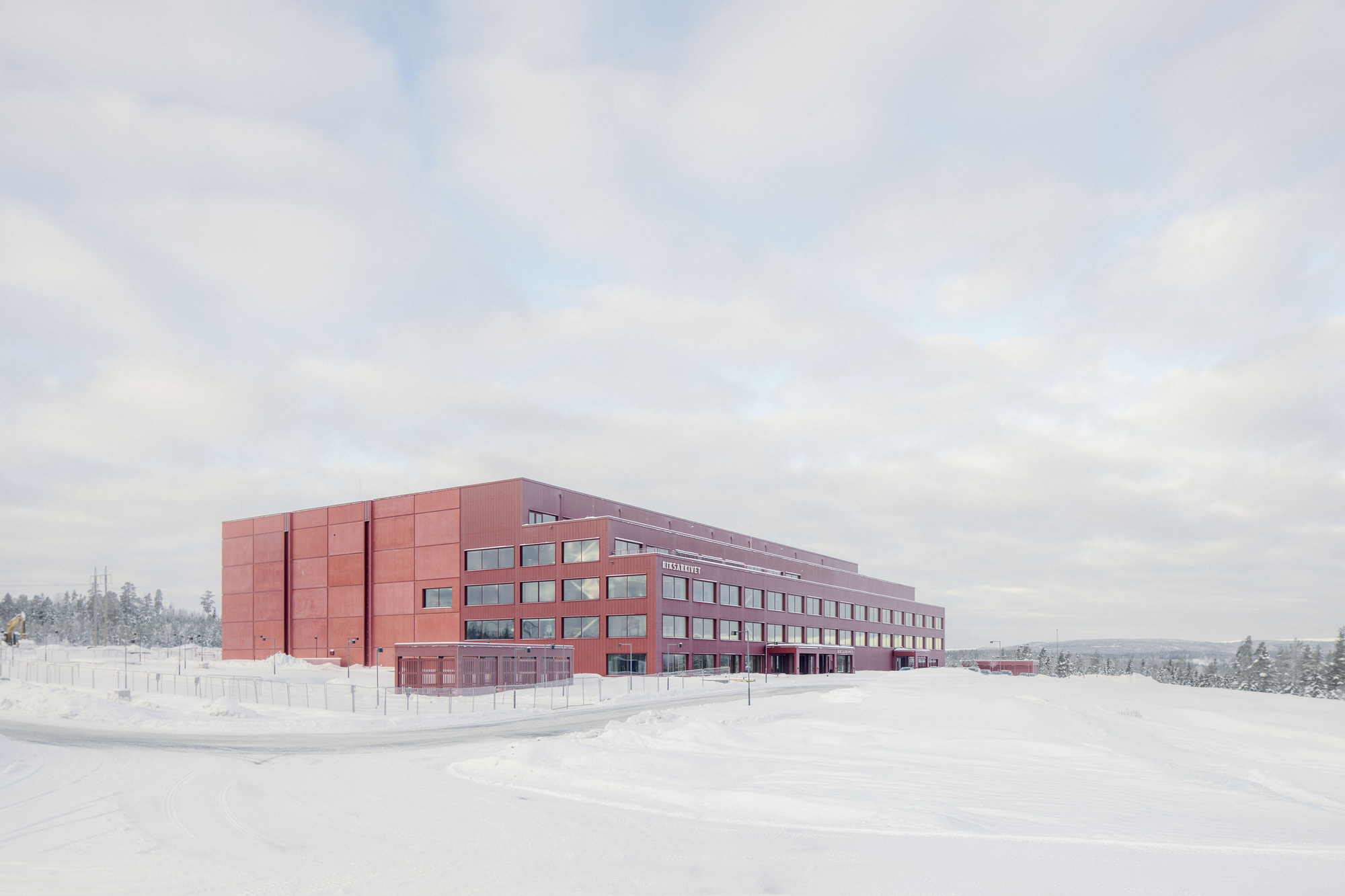 Four-storey red clad building with ribbon windows set back from a plowed road in deep snow under overcast sky