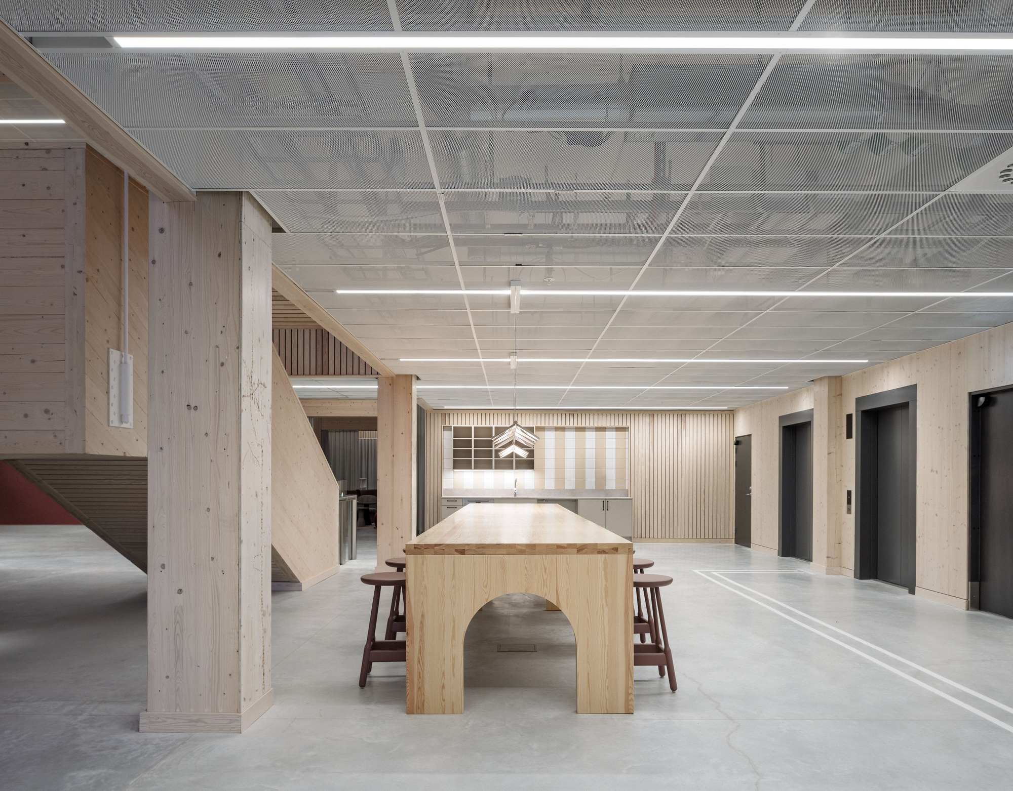 Ground floor hall with a long timber table with an arched base, glulam columns and lift doors under a mesh ceiling