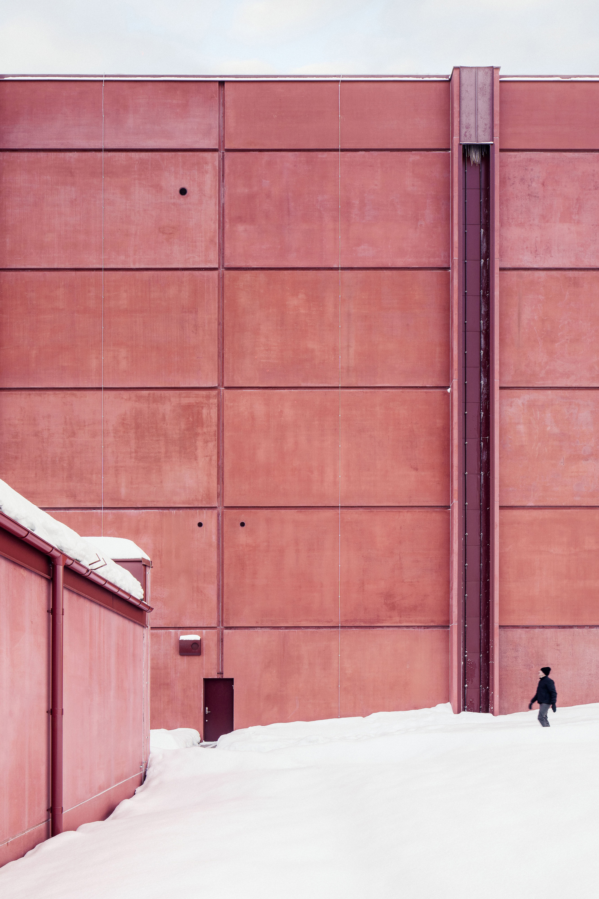 Blank red panelled wall with a vertical vent strip and small door, a lone pedestrian walking across the snow
