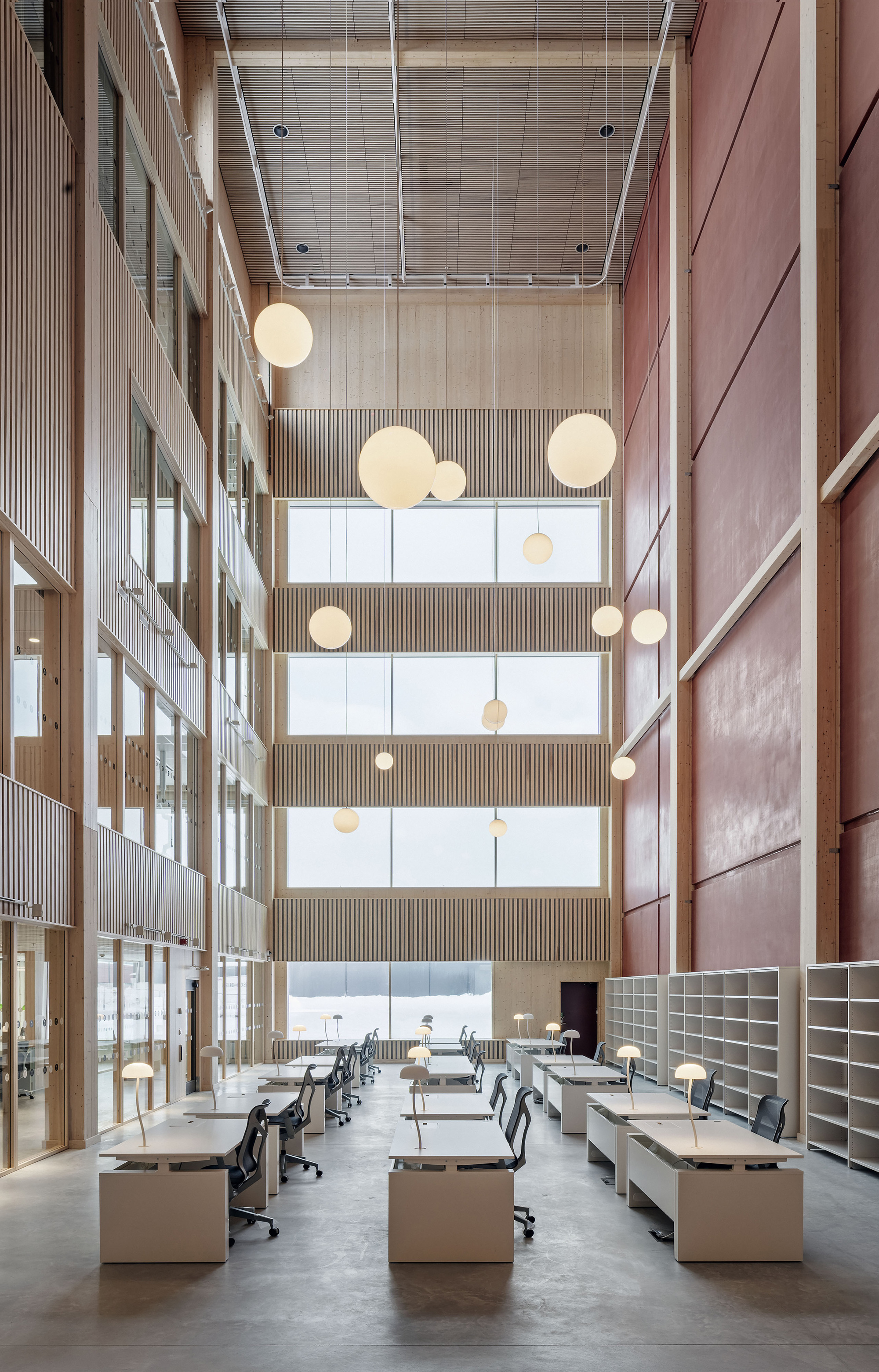 Tall reading hall with white desks, globe pendant lamps and shelving along a red wall, lit by winter daylight through stacked windows