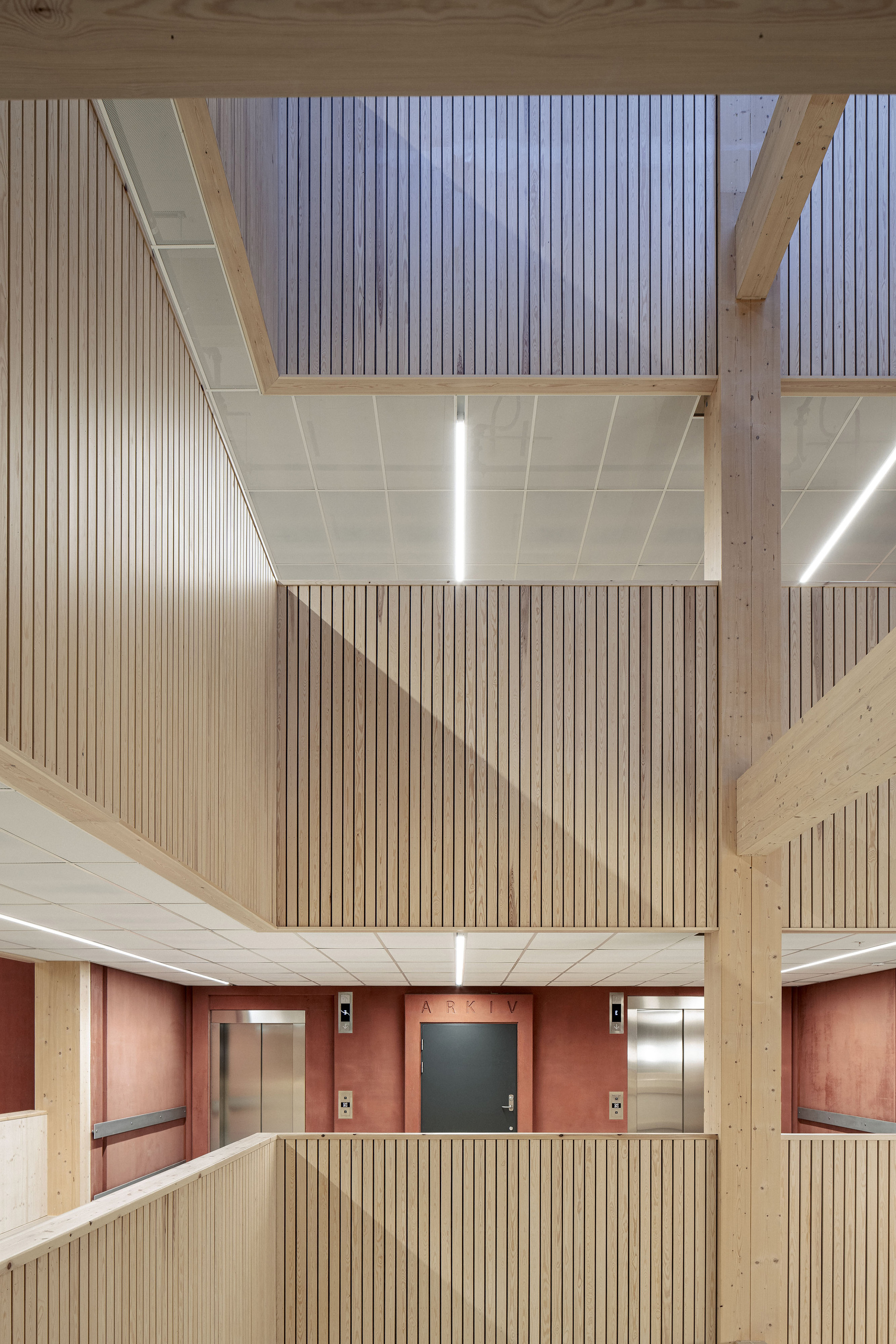 Timber atrium with slatted balustrades, glulam columns and beams, and a grey archive door set in a red wall