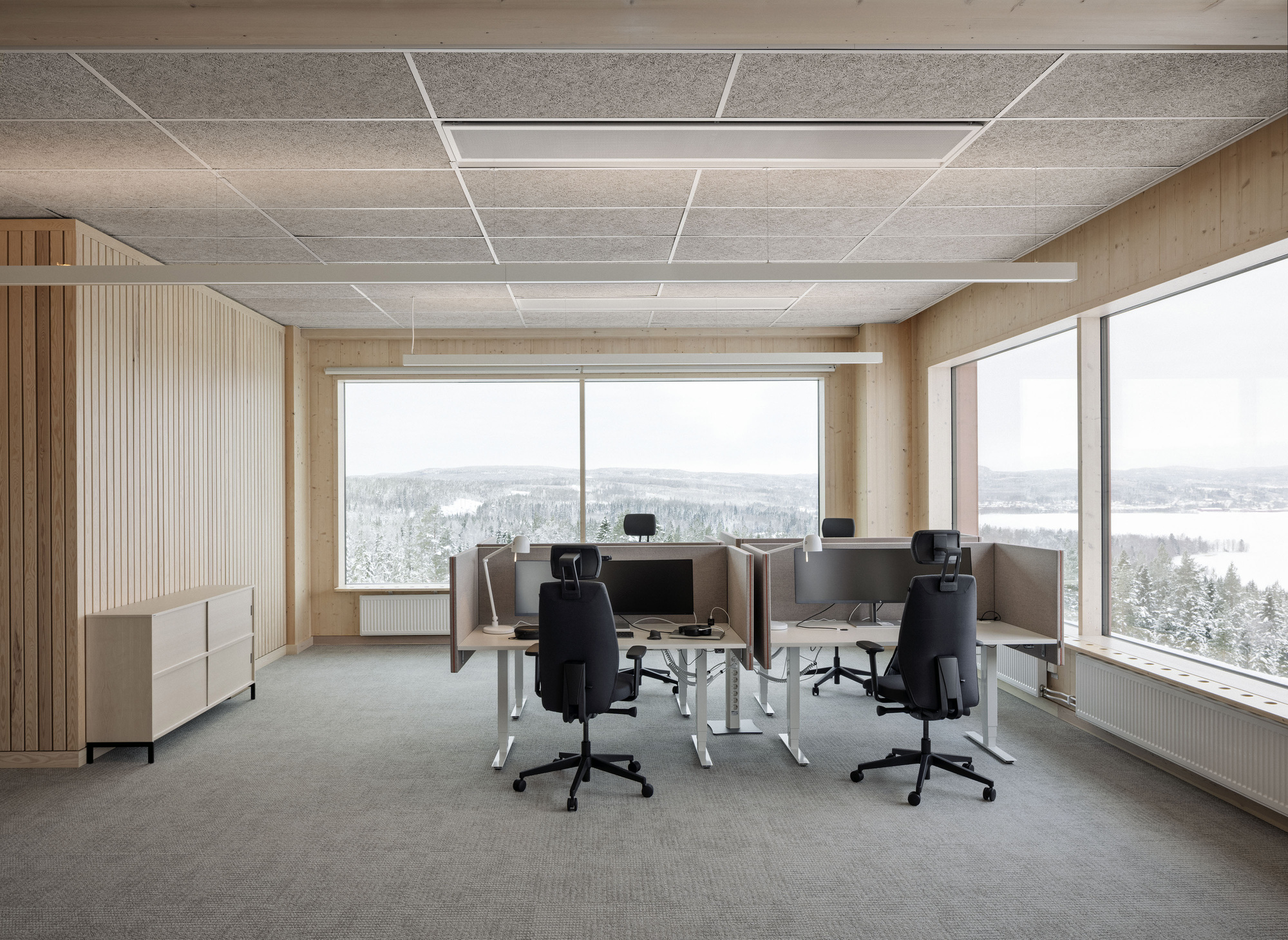 Open office with height-adjustable desks and black chairs facing corner windows onto a snowy forested valley and frozen lake