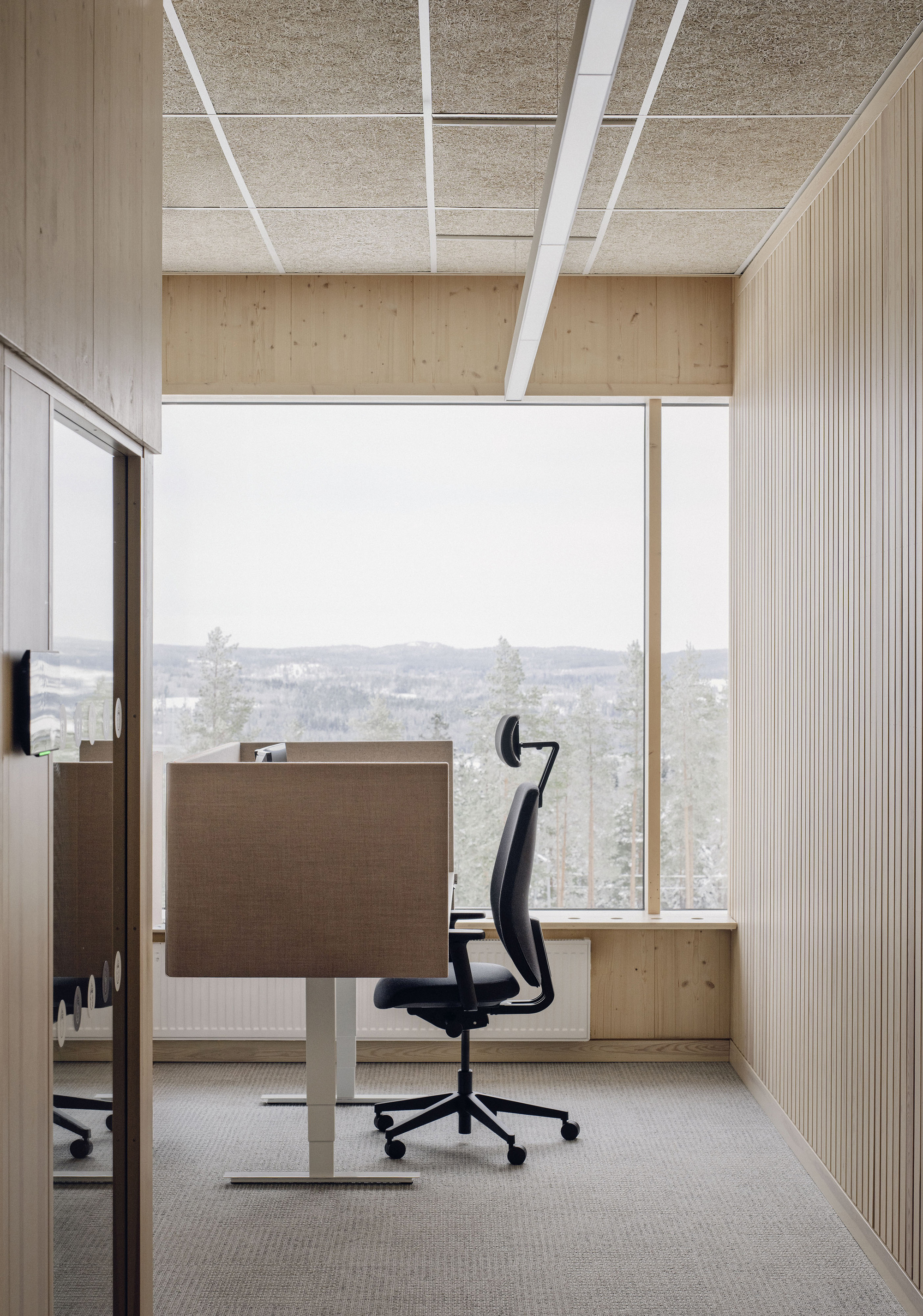 Single workstation with a black chair beside a slatted timber wall, looking through a window to snowy pines under a pale sky