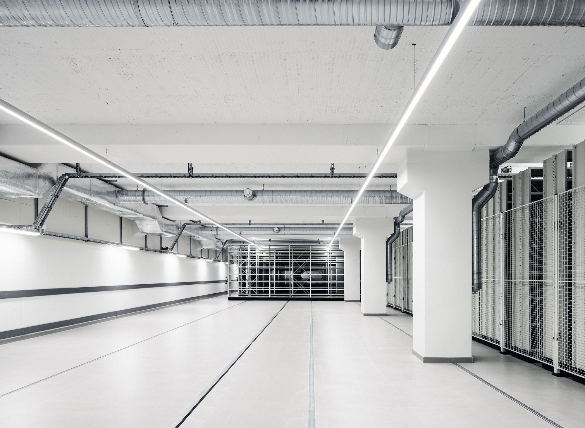 Basement archive corridor with exposed ductwork, linear ceiling lights, white columns, floor rails and mesh-fronted mobile shelving on the right