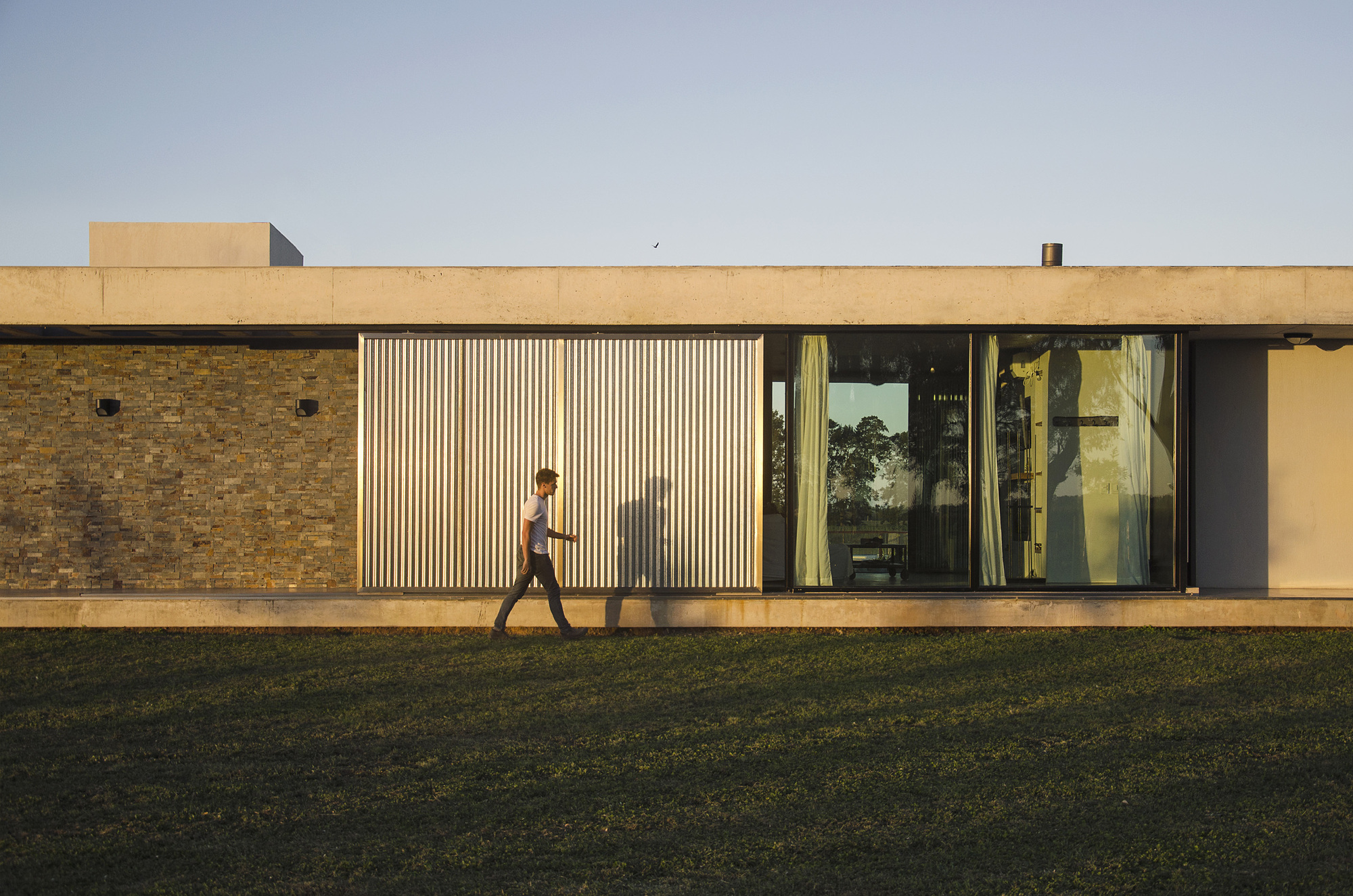 Side facade with stone wall, corrugated metal sliding panel and glazing under a concrete roof slab, a man walking past in low evening sun