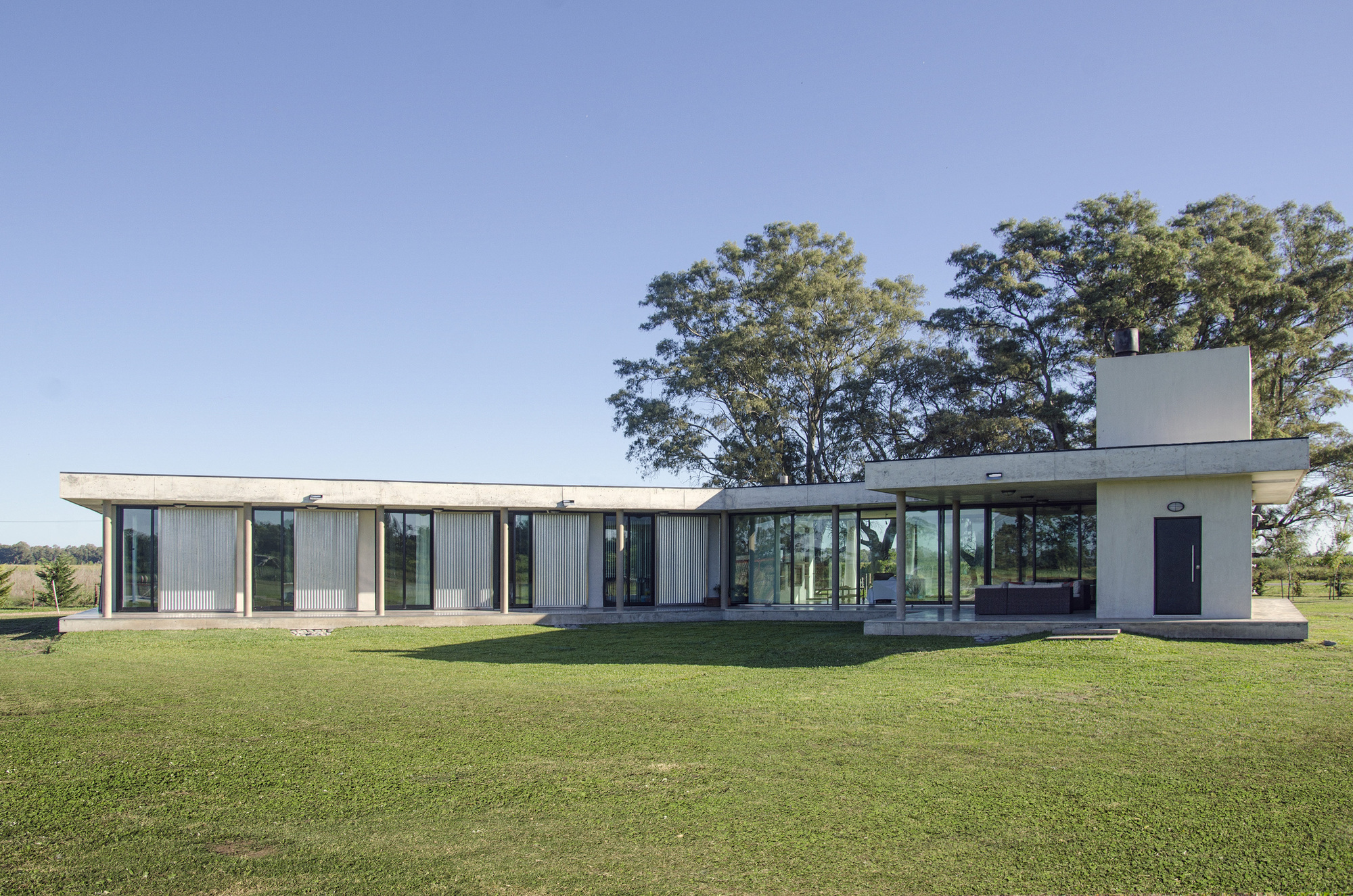L-shaped single-storey house with slim concrete columns and glass walls, large trees behind and a clear blue sky above the lawn