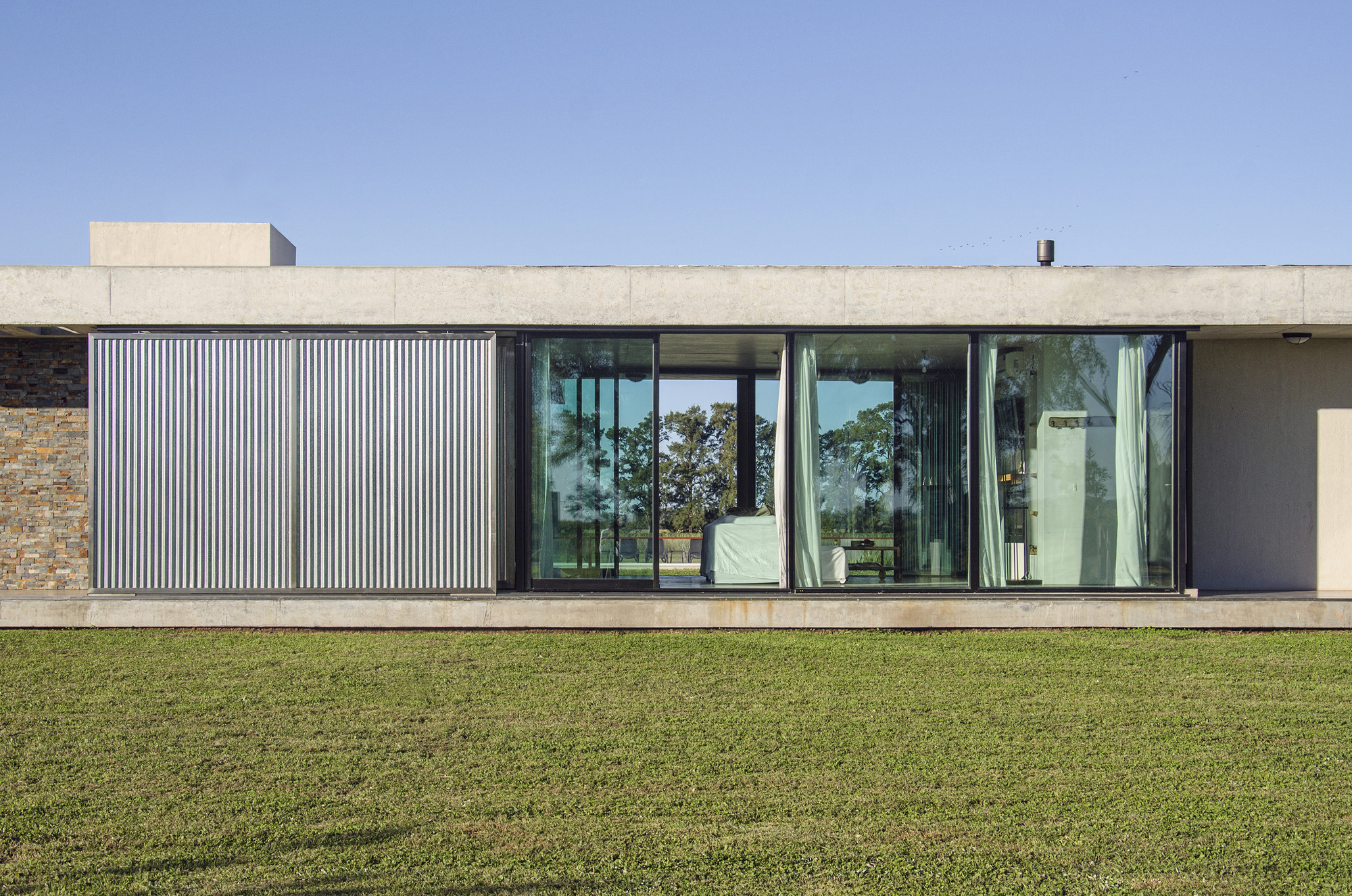 Frontal view of the corrugated metal shutter beside floor-to-ceiling glass sliders with pale curtains, on a green lawn in daylight