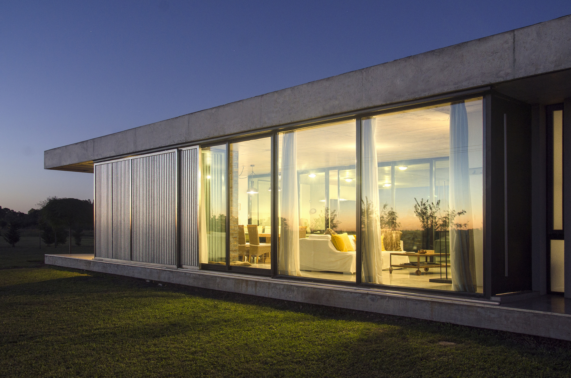 Corner of the glazed living room lit warmly from inside at dusk, with sliding metal louvers beside it and a deep blue sky