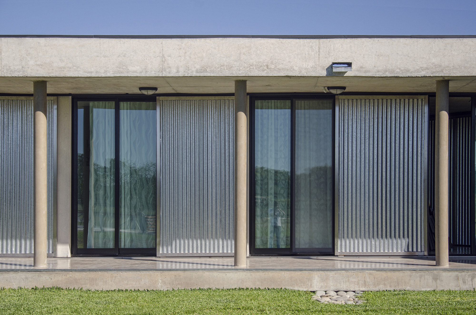 Covered porch with round concrete columns, black-framed glass doors and corrugated translucent panels, a few river stones set in the grass