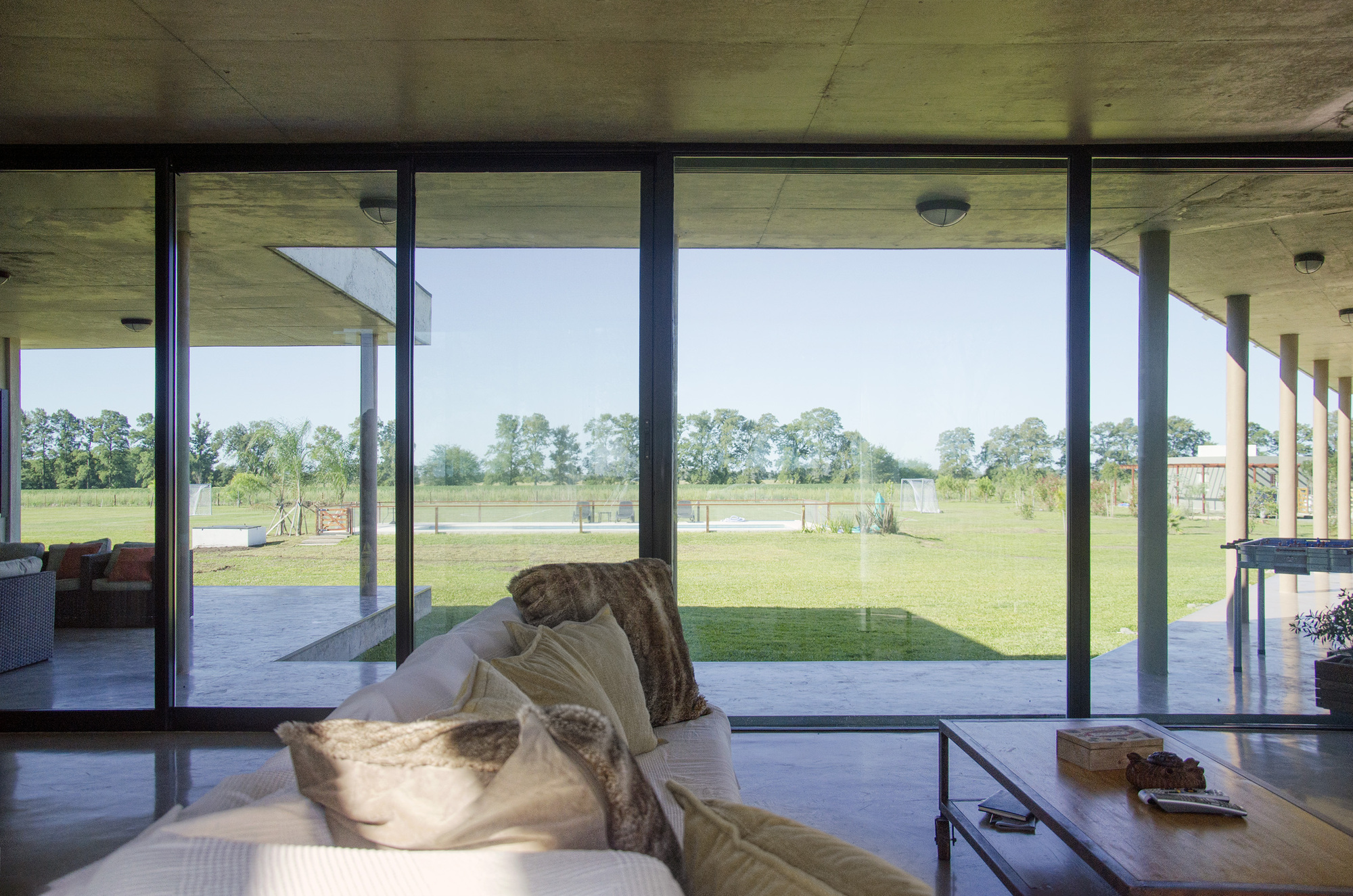 Interior view from a sofa with fur cushions through sliding glass walls to the covered terrace, open lawn and a line of trees