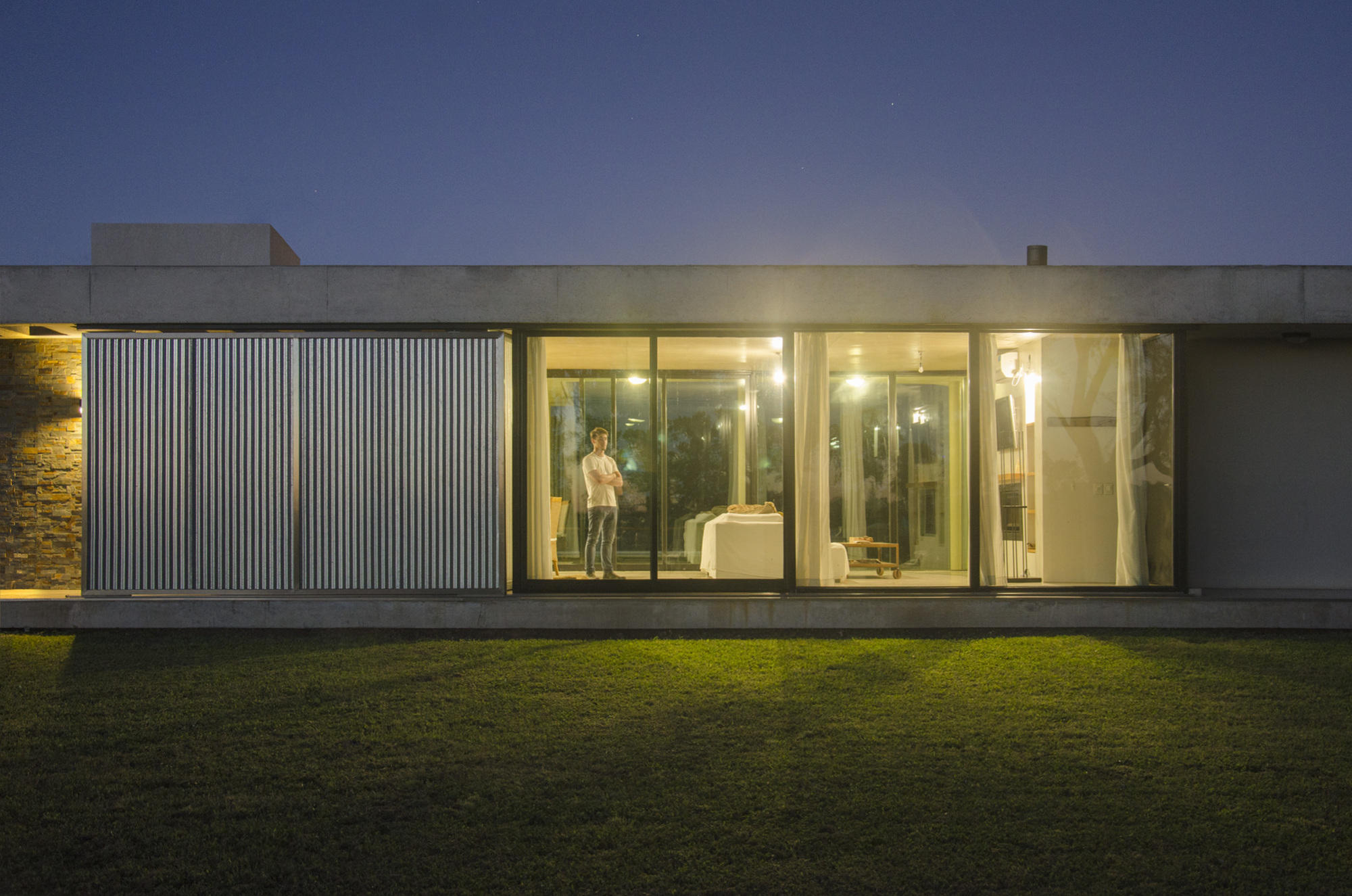 Glazed front of the single-storey house at dusk with corrugated metal shutters and a person standing inside the lit room