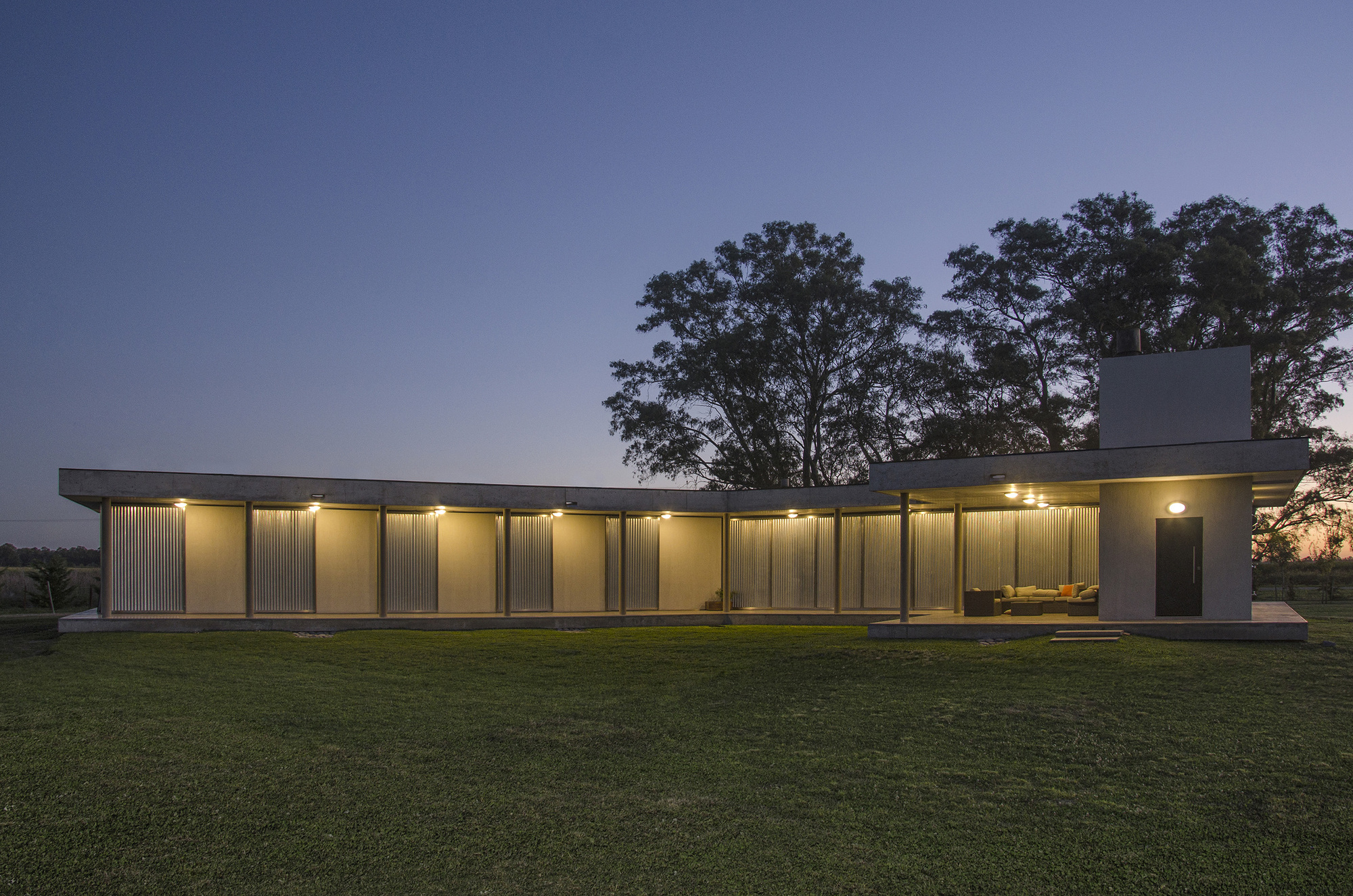 Long L-shaped concrete pavilion with slim columns and corrugated screens, lit from within at dusk beside a tall eucalyptus