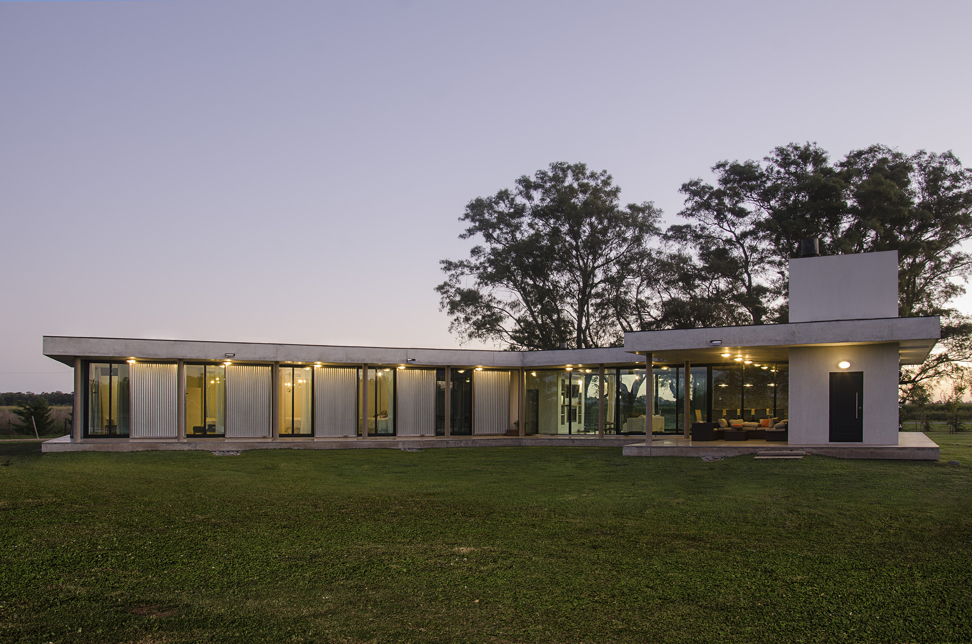 Wide view of the flat-roofed house at twilight, glazed walls glowing behind columns, with a covered porch and chimney at right