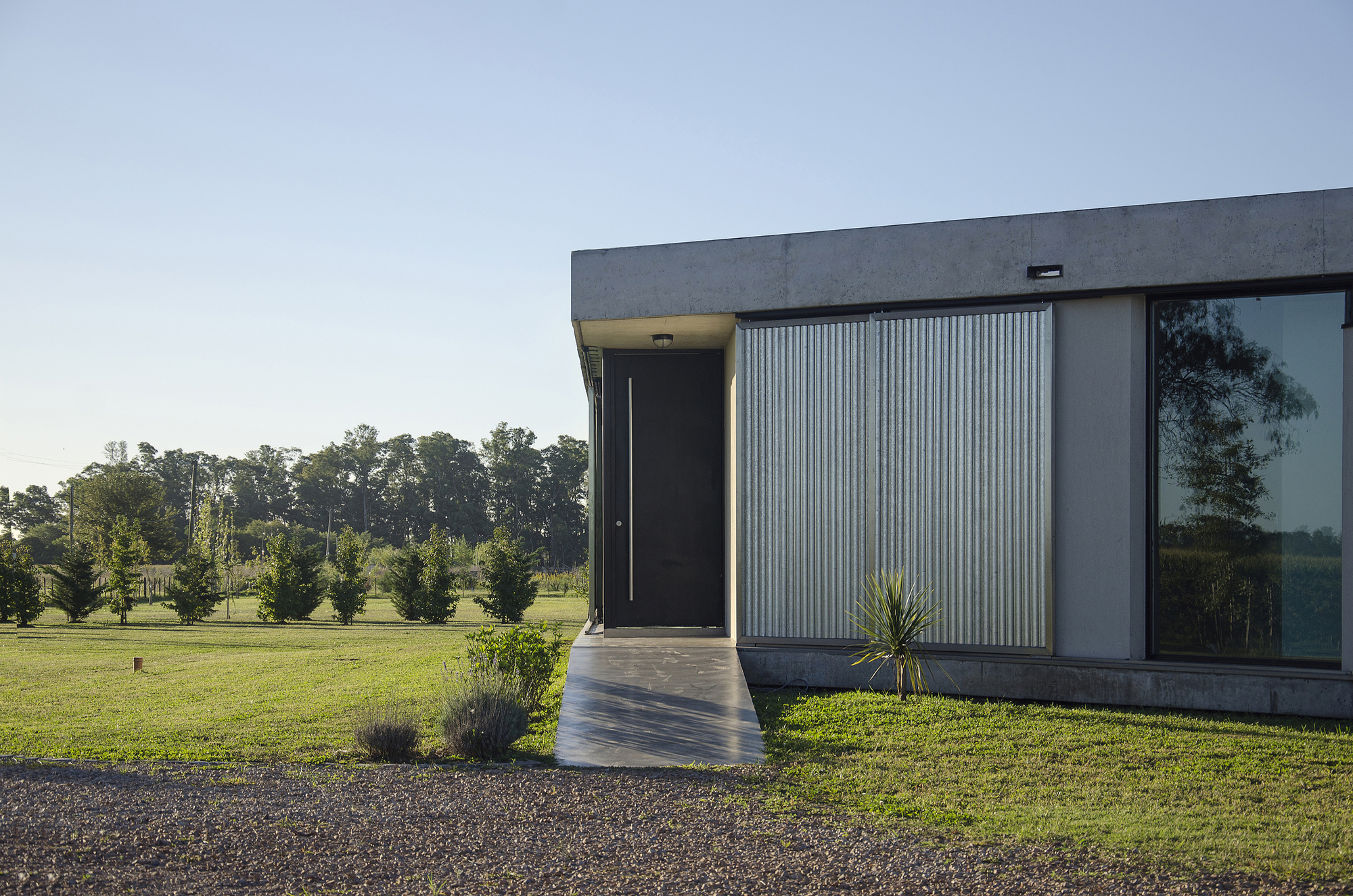 Black entry door at the end of a concrete path beside corrugated metal shutters, with a yucca plant and gravel in afternoon light