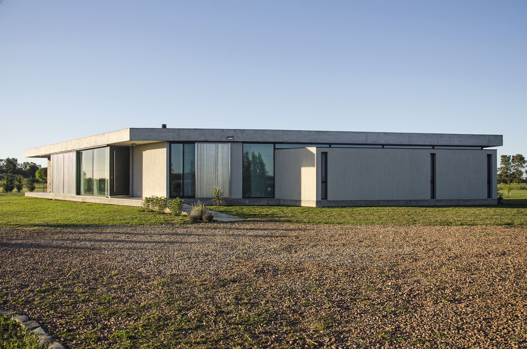 Low concrete house with a flat cantilevered roof, corrugated metal panels and tall glazing, seen across a gravel drive in late afternoon sun
