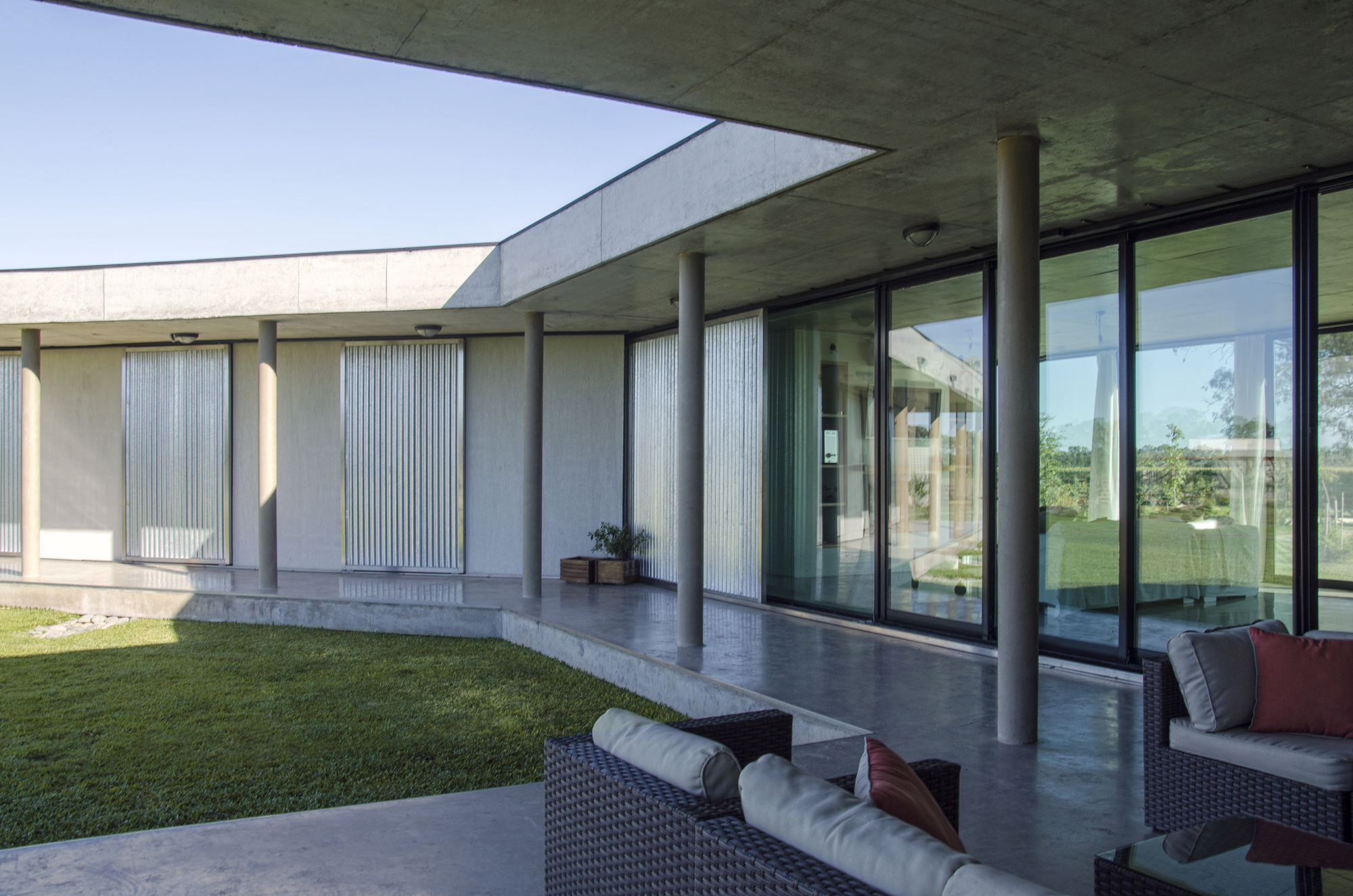 Covered concrete porch with slender round columns facing a grass courtyard, wicker seating in the foreground and sliding glass doors at right