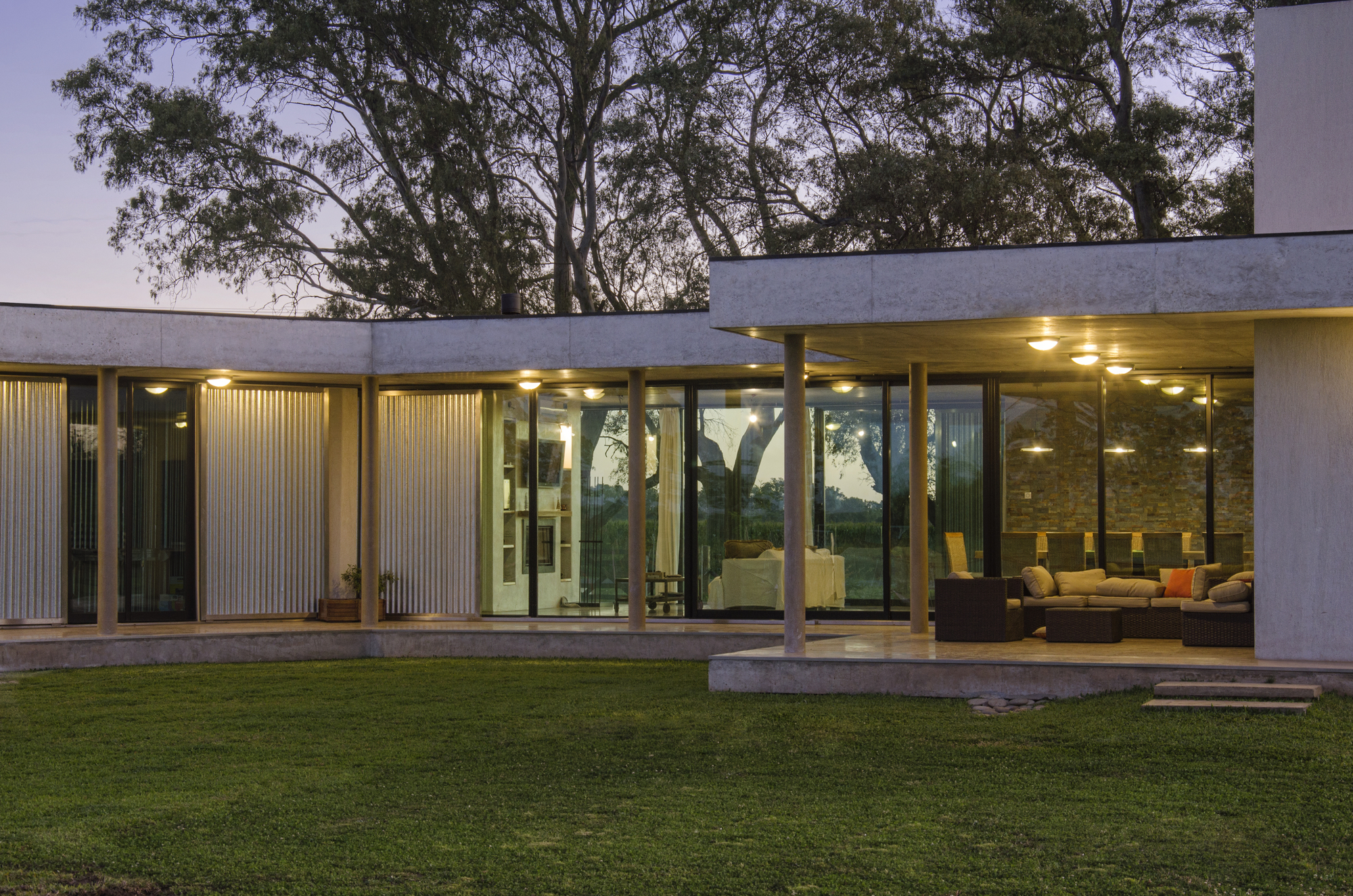 Courtyard facade at dusk with lit ceiling lamps, slim columns and glazed walls reflecting eucalyptus trees above a lawn