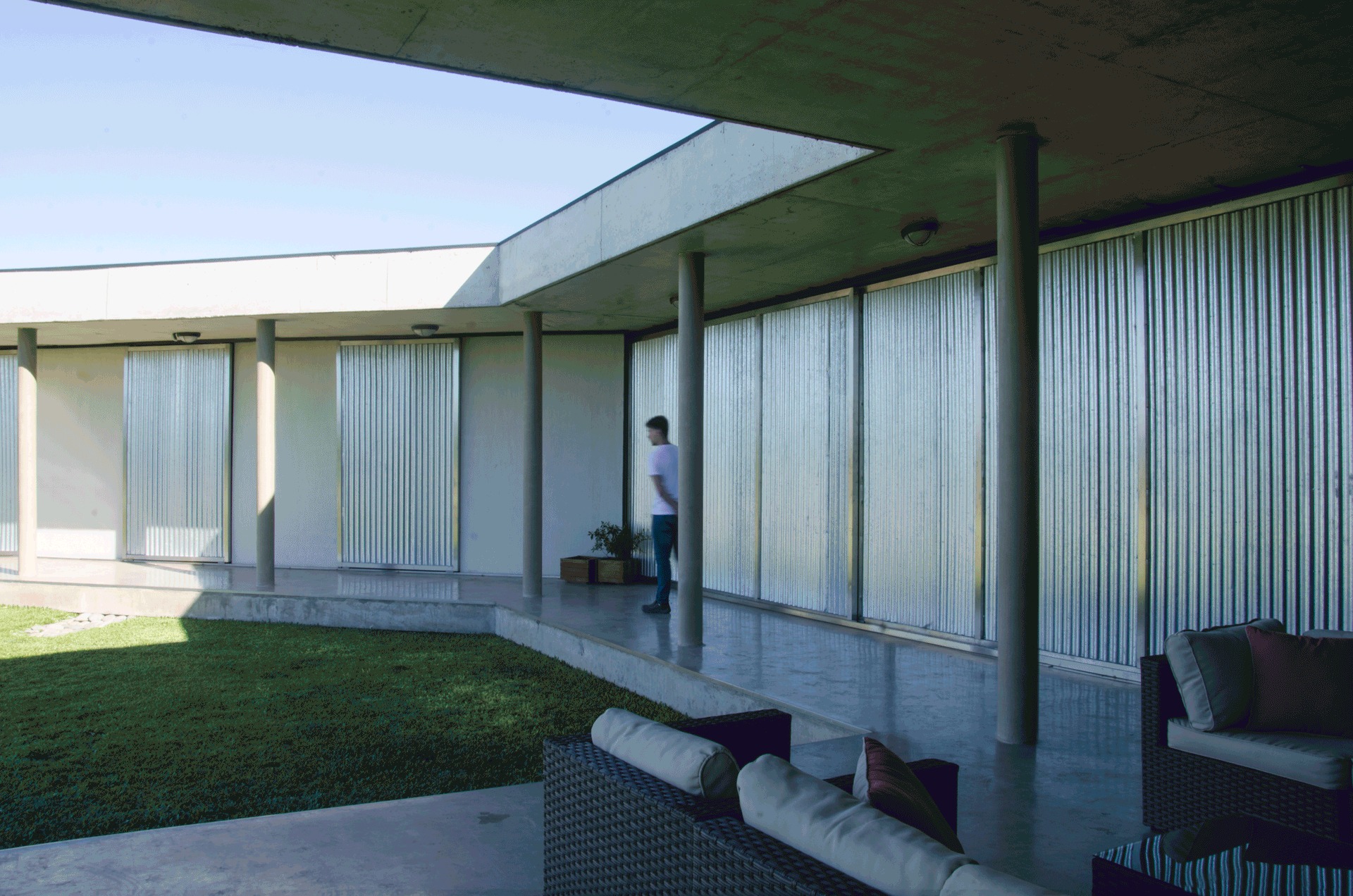 Person standing on the concrete porch beside corrugated metal sliding shutters, with columns and a grass courtyard in afternoon light
